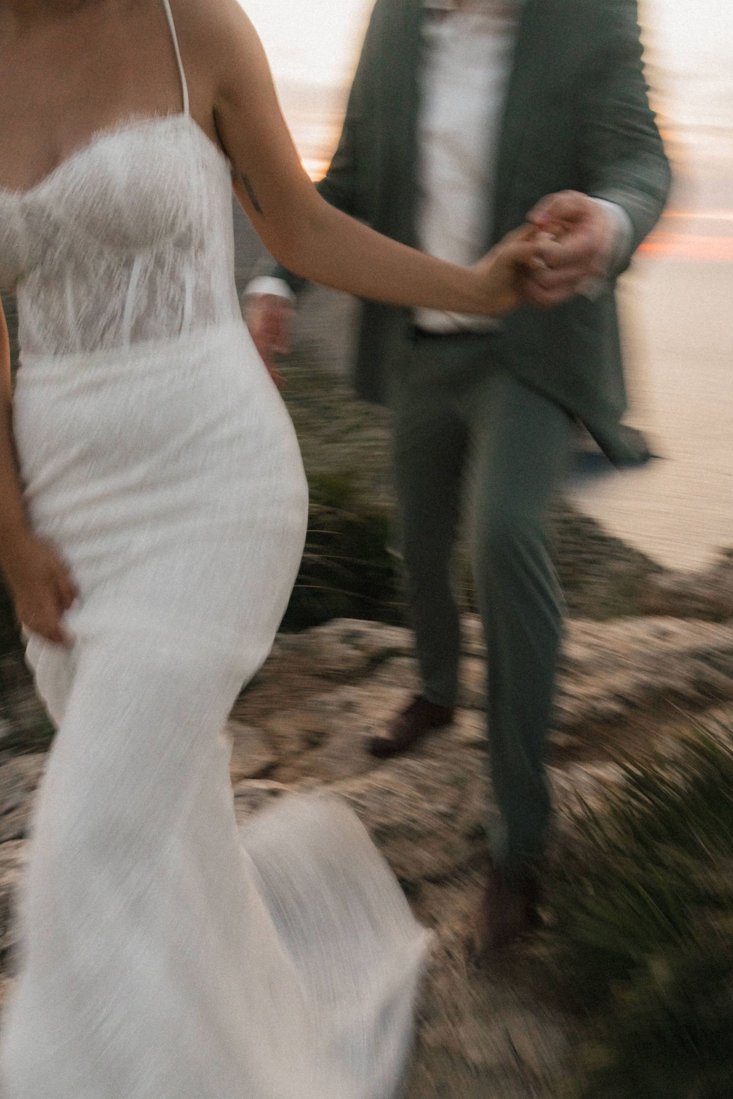 Bride in white lace gown and groom in gray suit hold hands while walking on rocky terrain at dusk.