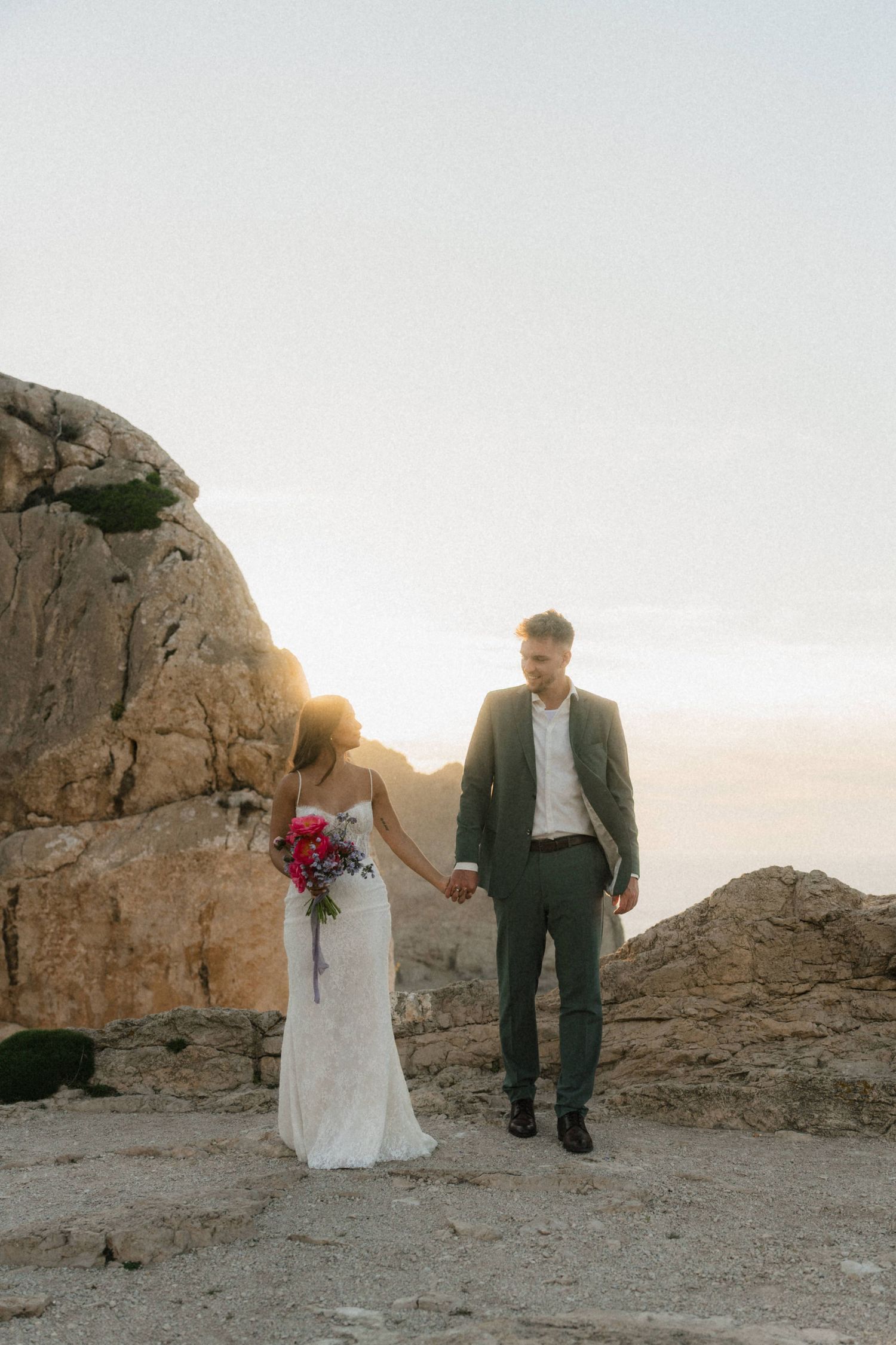 Bride and groom holding hands at sunset on rocky cliffs, bride in white gown with red bouquet, groom in gray suit.