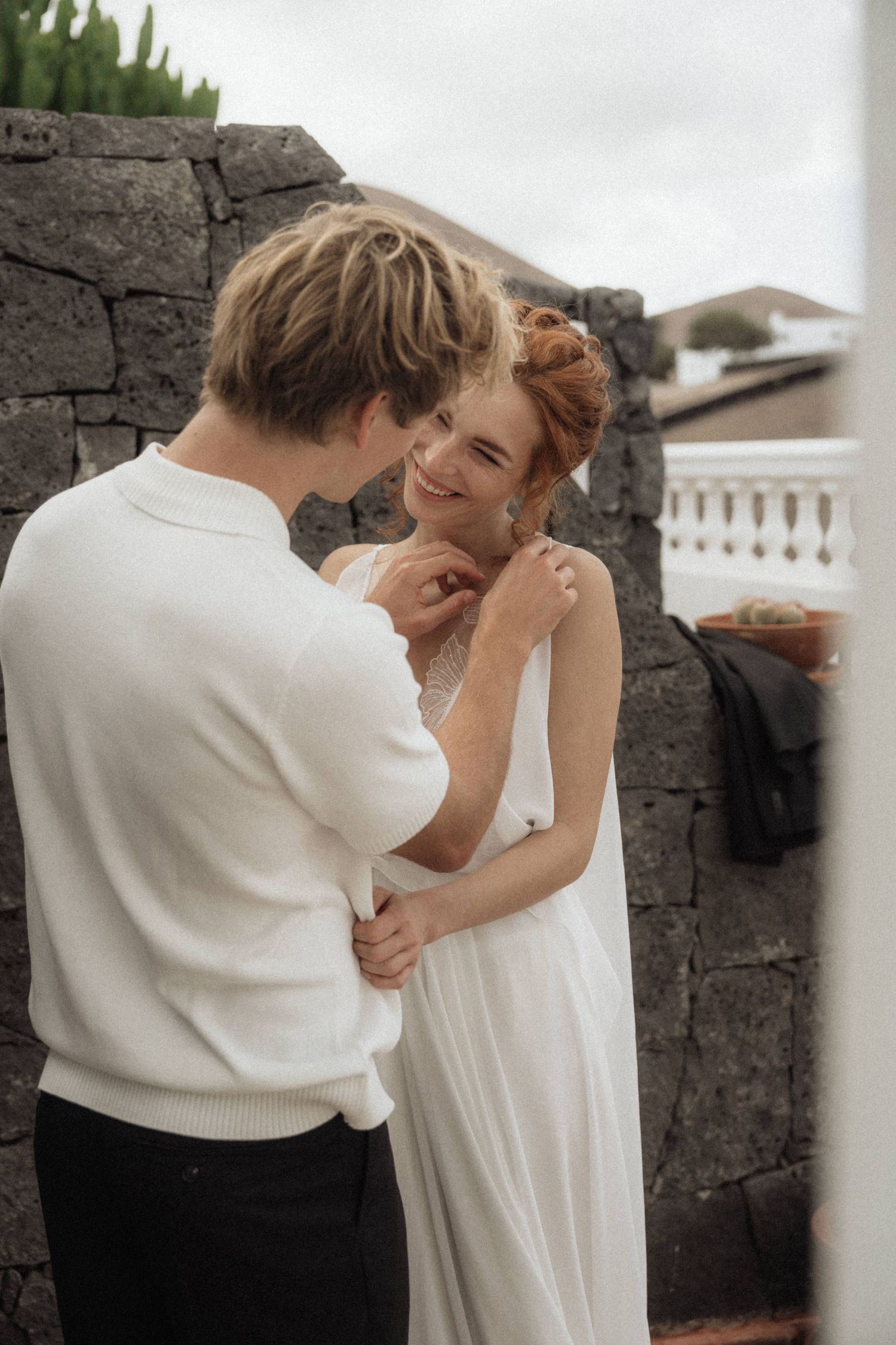 Smiling bride in white dress embraces groom in white sweater outdoors near stone wall.