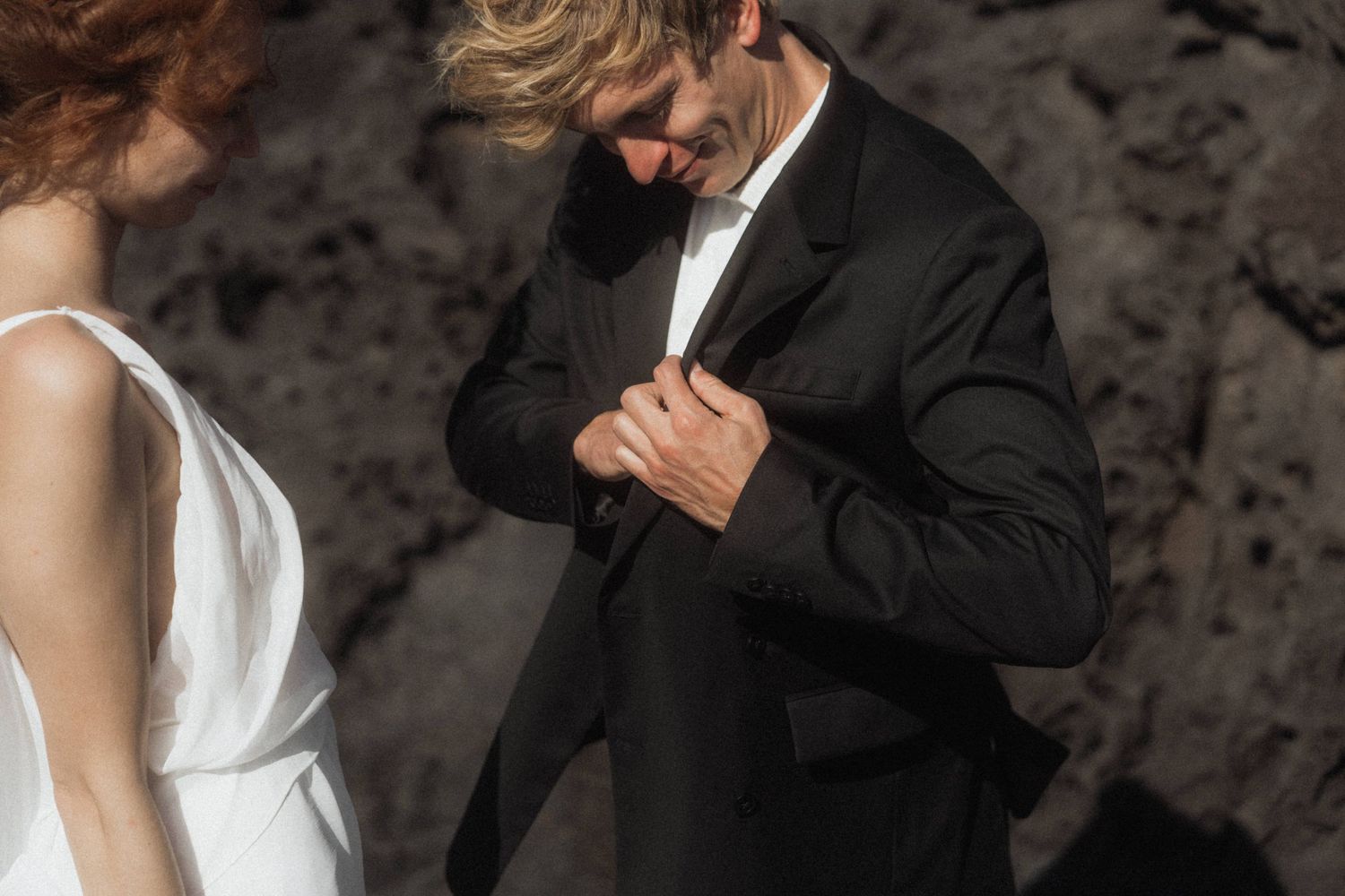 Groom in black suit adjusts jacket beside bride in white dress at rocky backdrop.