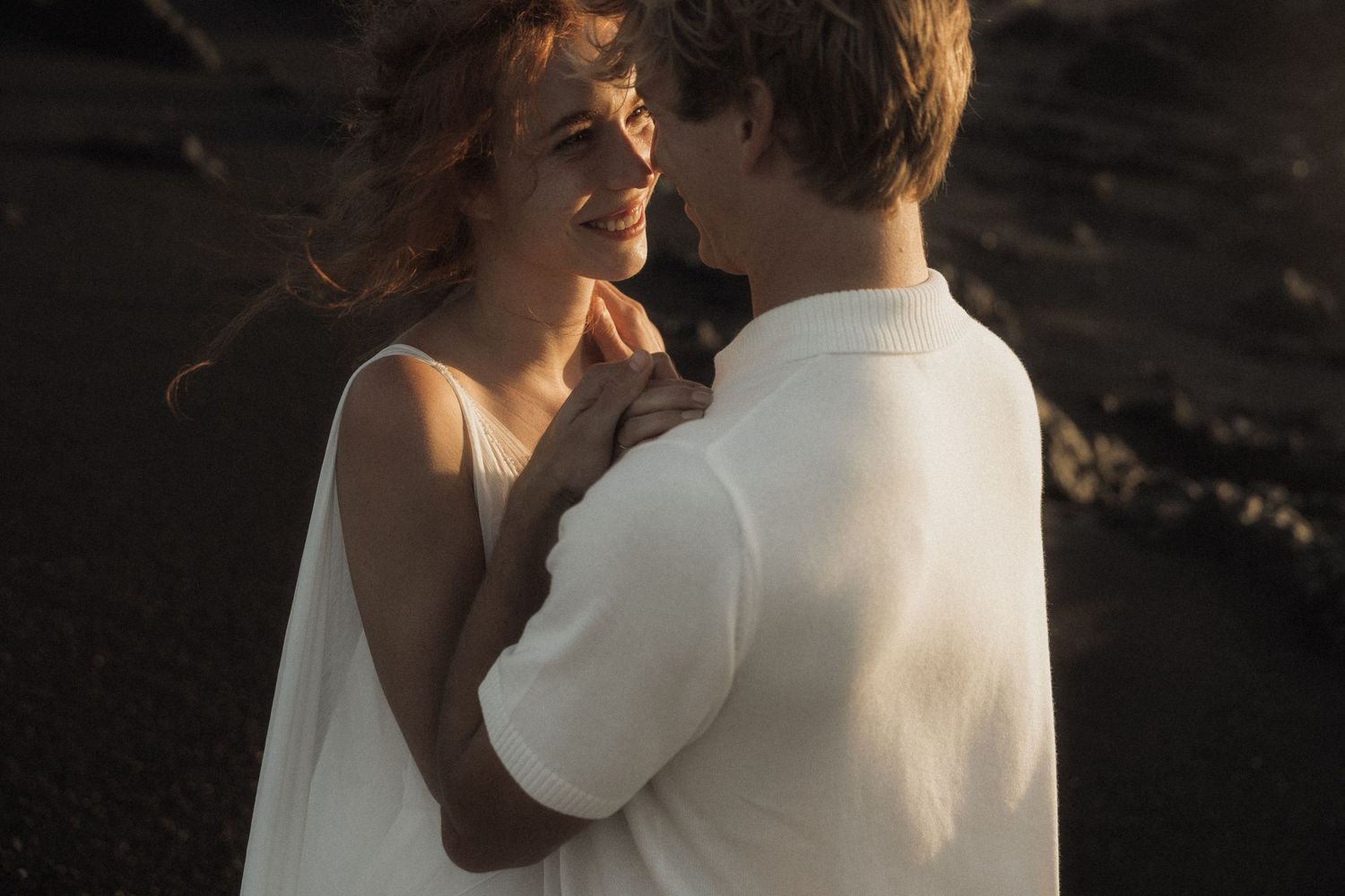 Couple sharing romantic moment at golden sunset on dark sand beach in white attire.