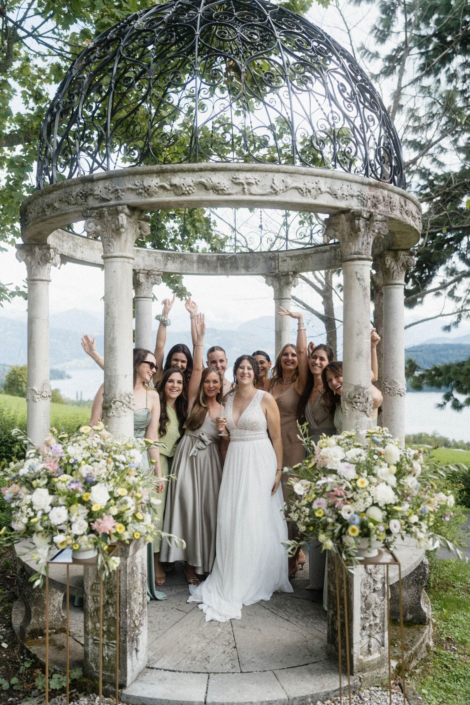 Bride in white gown with bridesmaids in sage and taupe dresses celebrating under an ornate stone gazebo with lake views.