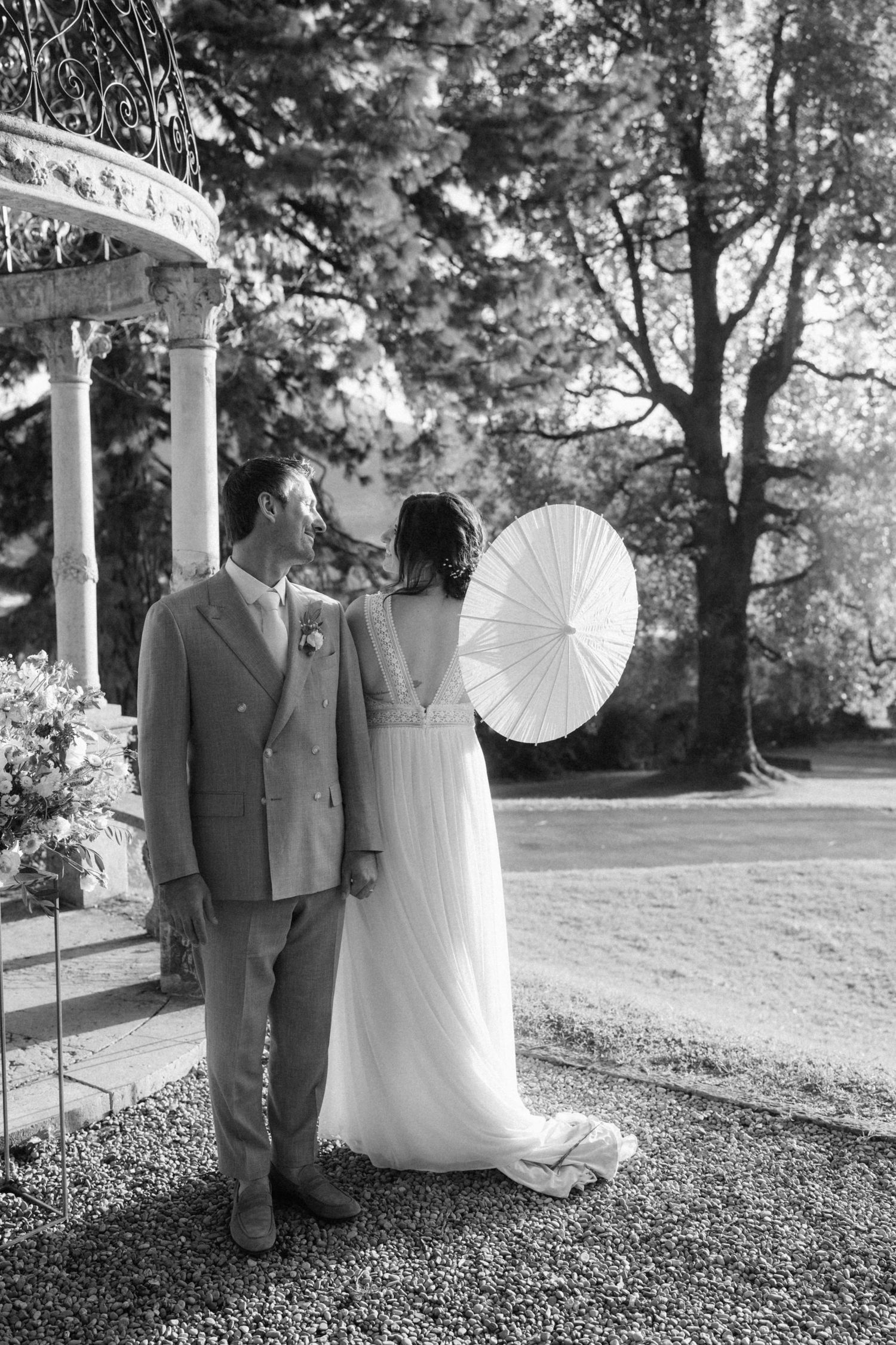 Black and white photo of bride and groom standing near garden gazebo, bride holding white parasol umbrella.