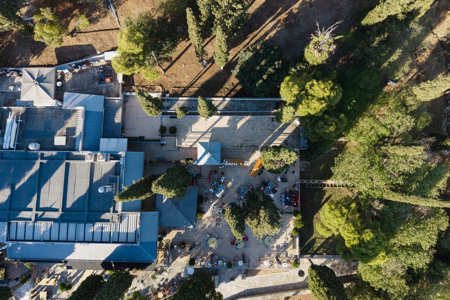 Aerial drone view of a building complex surrounded by lush green trees and a paved courtyard area.