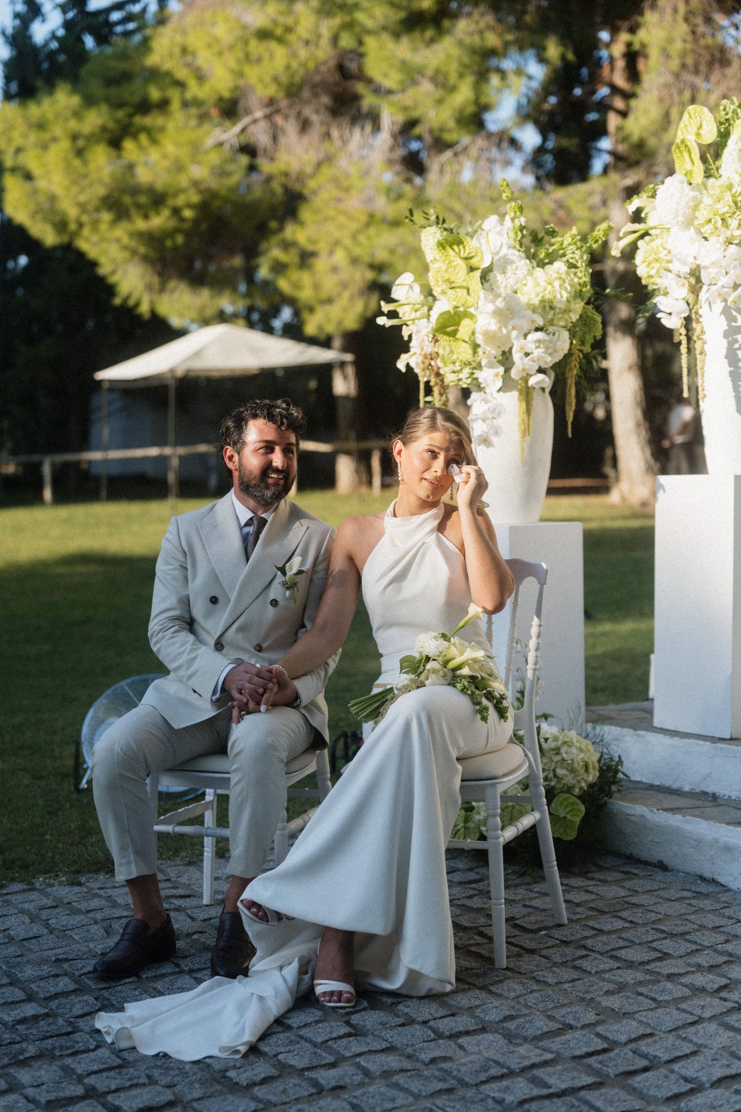 Bride and groom seated on white chairs at outdoor wedding ceremony with white floral arrangements.