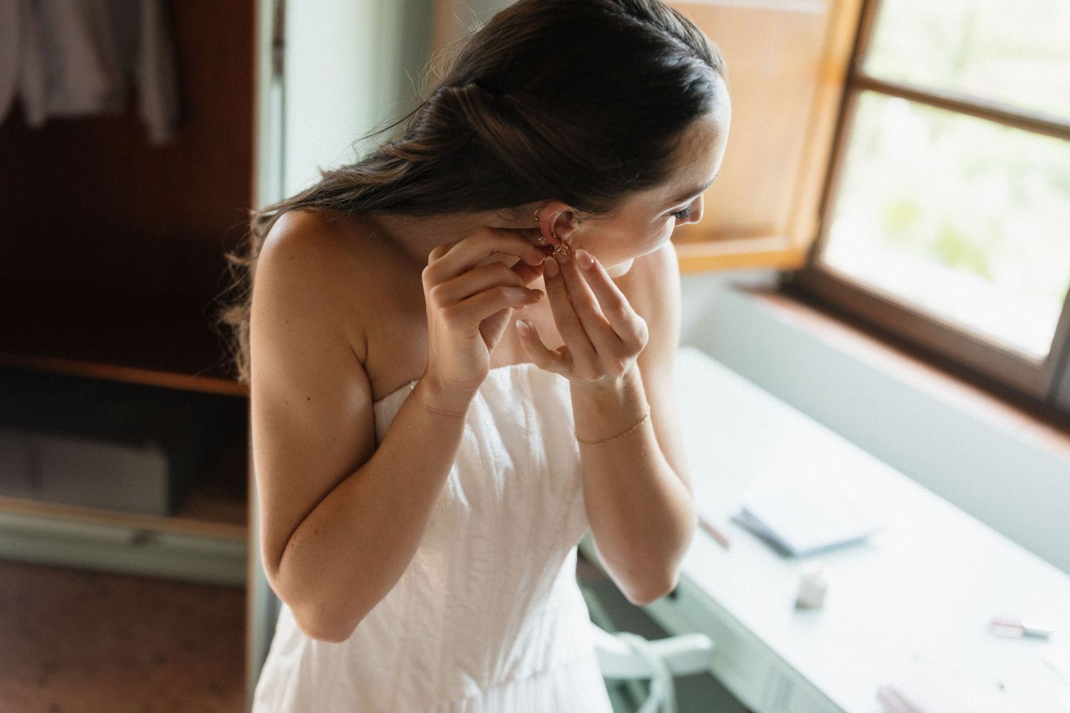 Bride in a white dress putting on an earring beside a sunlit window while getting ready on her wedding day.
