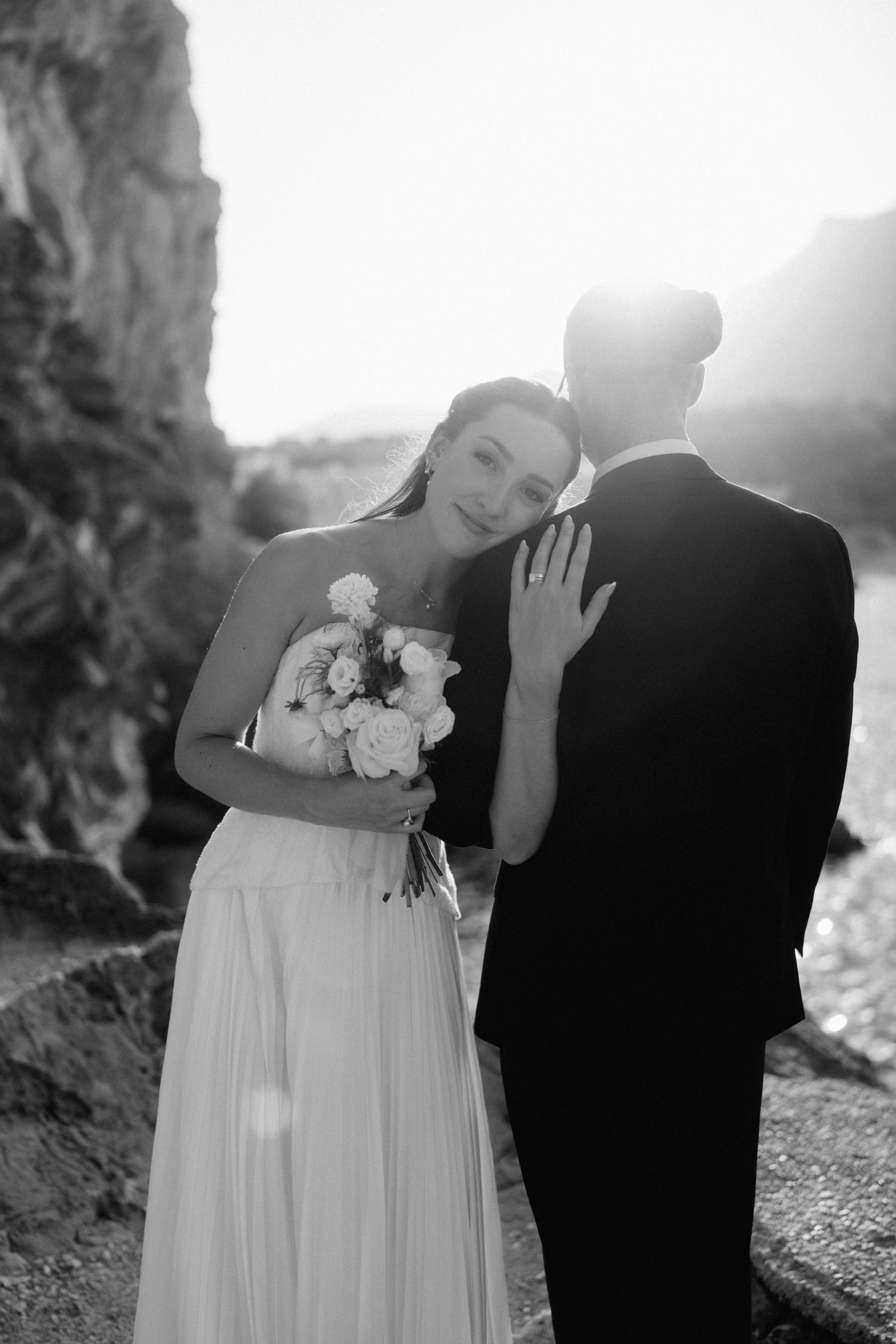 Black and white portrait of a bride holding a bouquet and resting her head on her groom's shoulder in warm backlight.