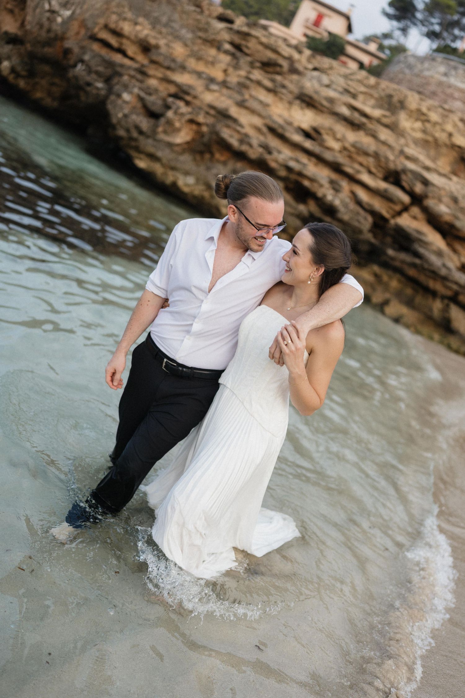 Couple in white wedding attire wading barefoot in shallow seawater, embracing near rocky cliffs during a beach wedding photoshoot.