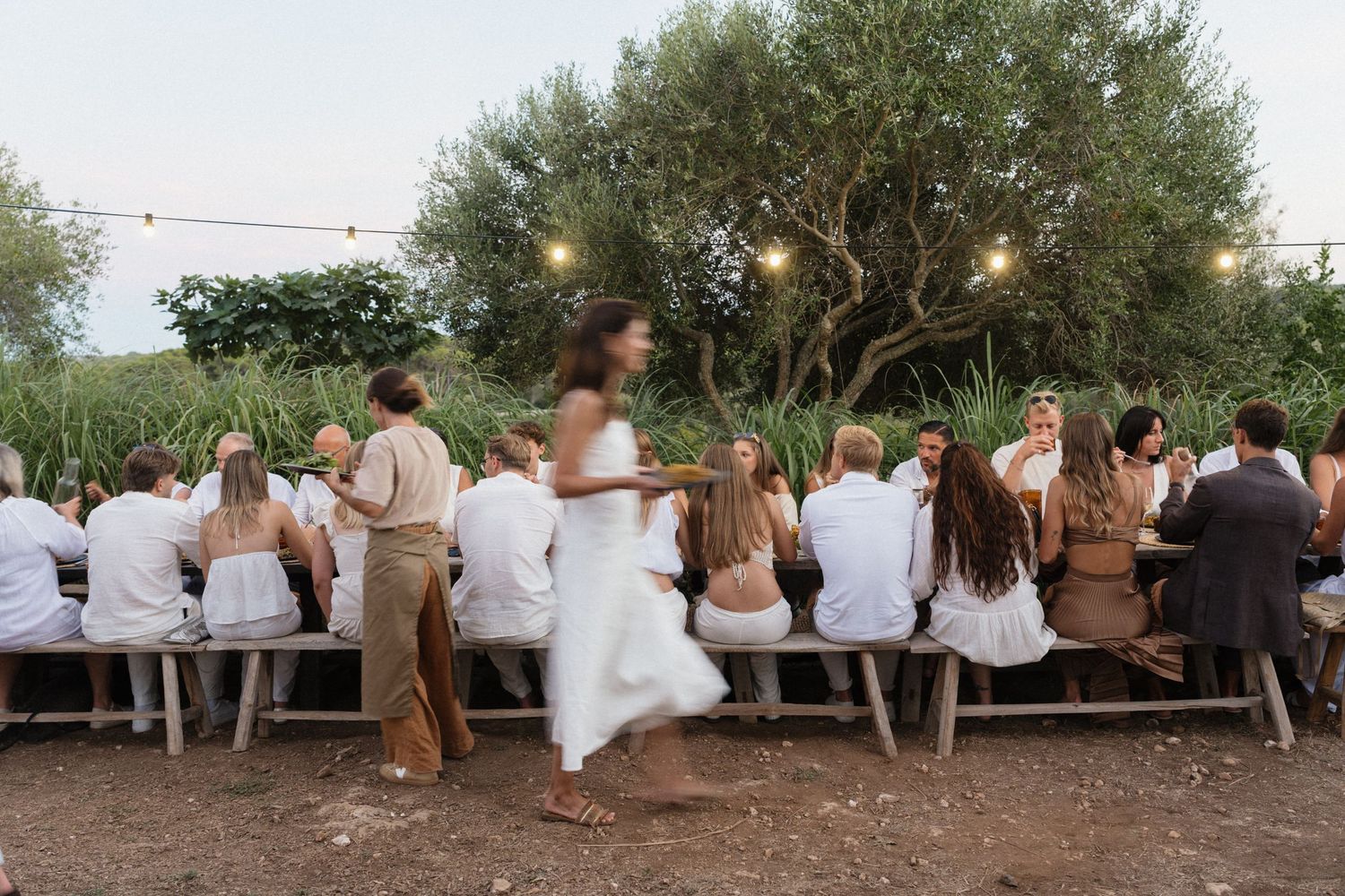Guests in white and neutral outfits sit at a long outdoor table under string lights as a woman in a white dress walks past at dusk.