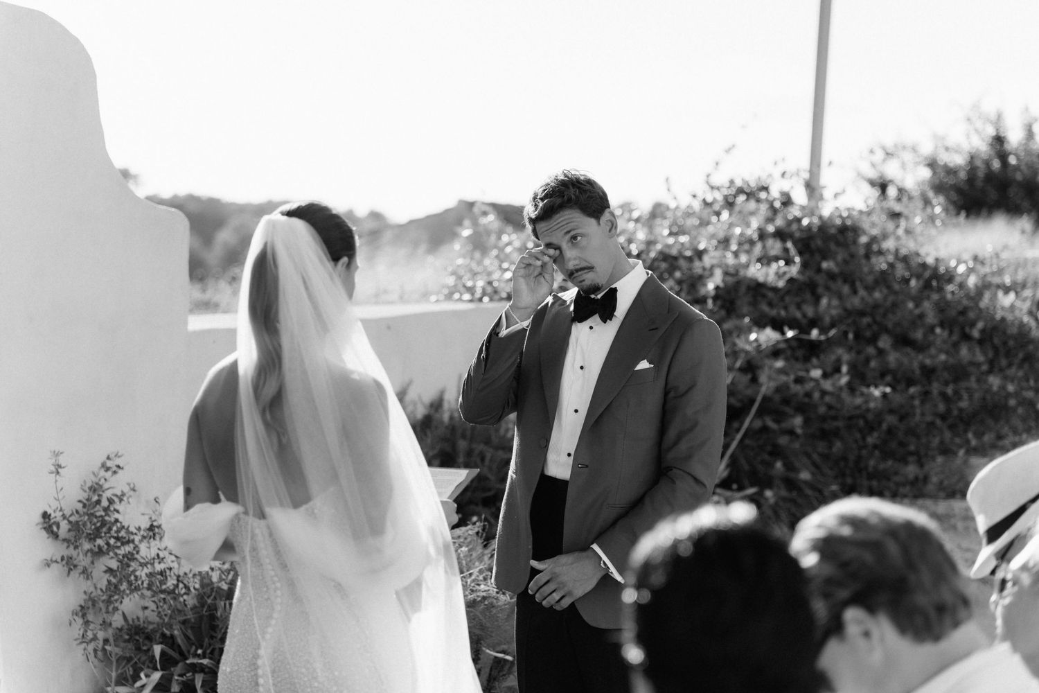 A groom in a dark suit and bow tie wipes his eye during an outdoor wedding ceremony as the veiled bride stands beside him.