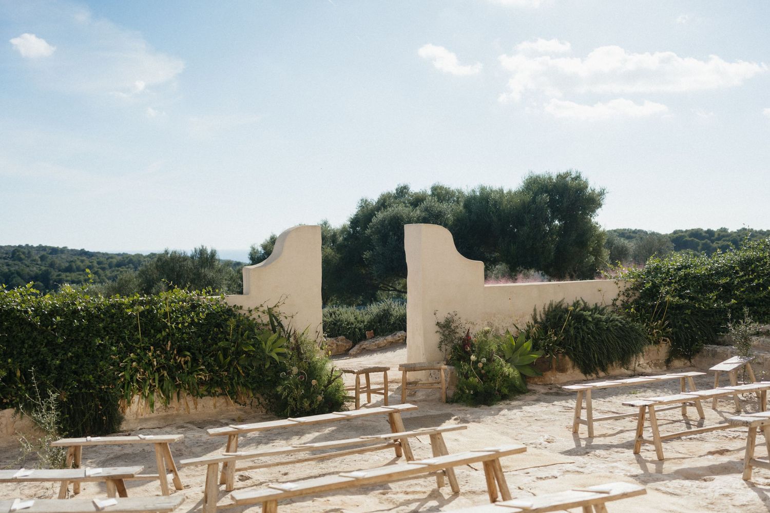 Outdoor wedding ceremony space with rustic wooden benches facing stone walls, olive trees, and lush greenery under a soft blue sky.