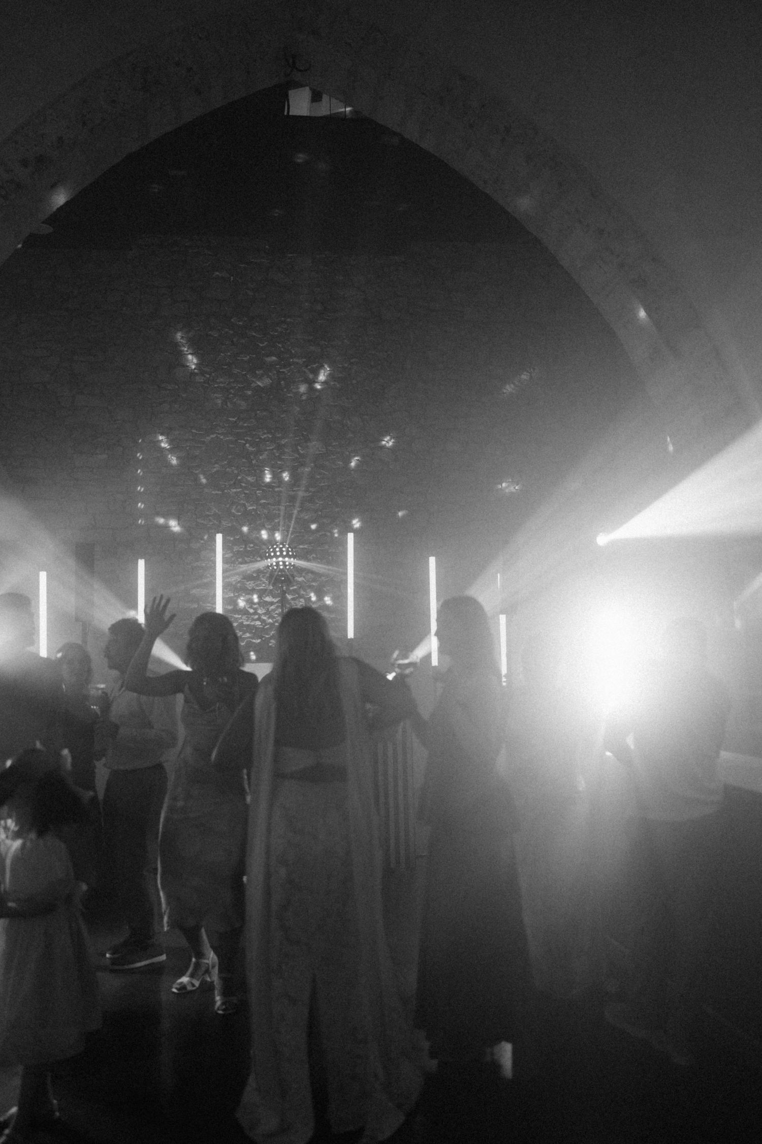 Black and white photo of silhouetted guests dancing under a stone arch with glowing lights at an elegant event.