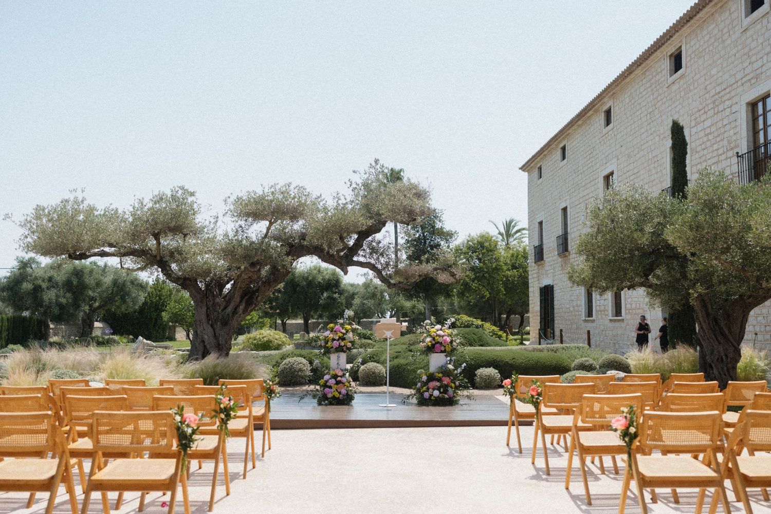 Outdoor wedding ceremony setup with wooden chairs, olive trees, and floral arrangements at a rustic Mediterranean venue.