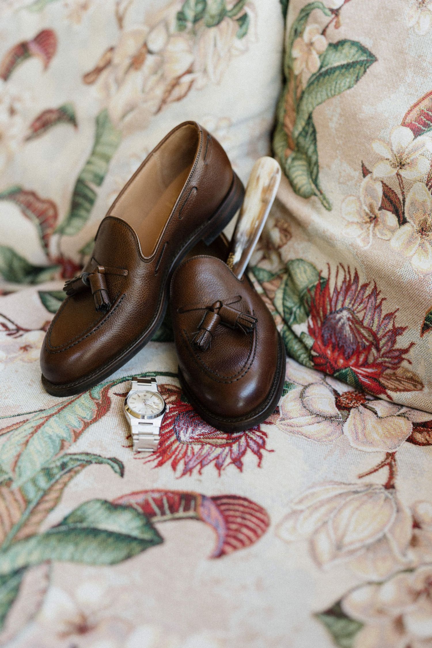 Brown leather tassel loafers with a gold watch on floral fabric, perfect wedding day groom accessories.