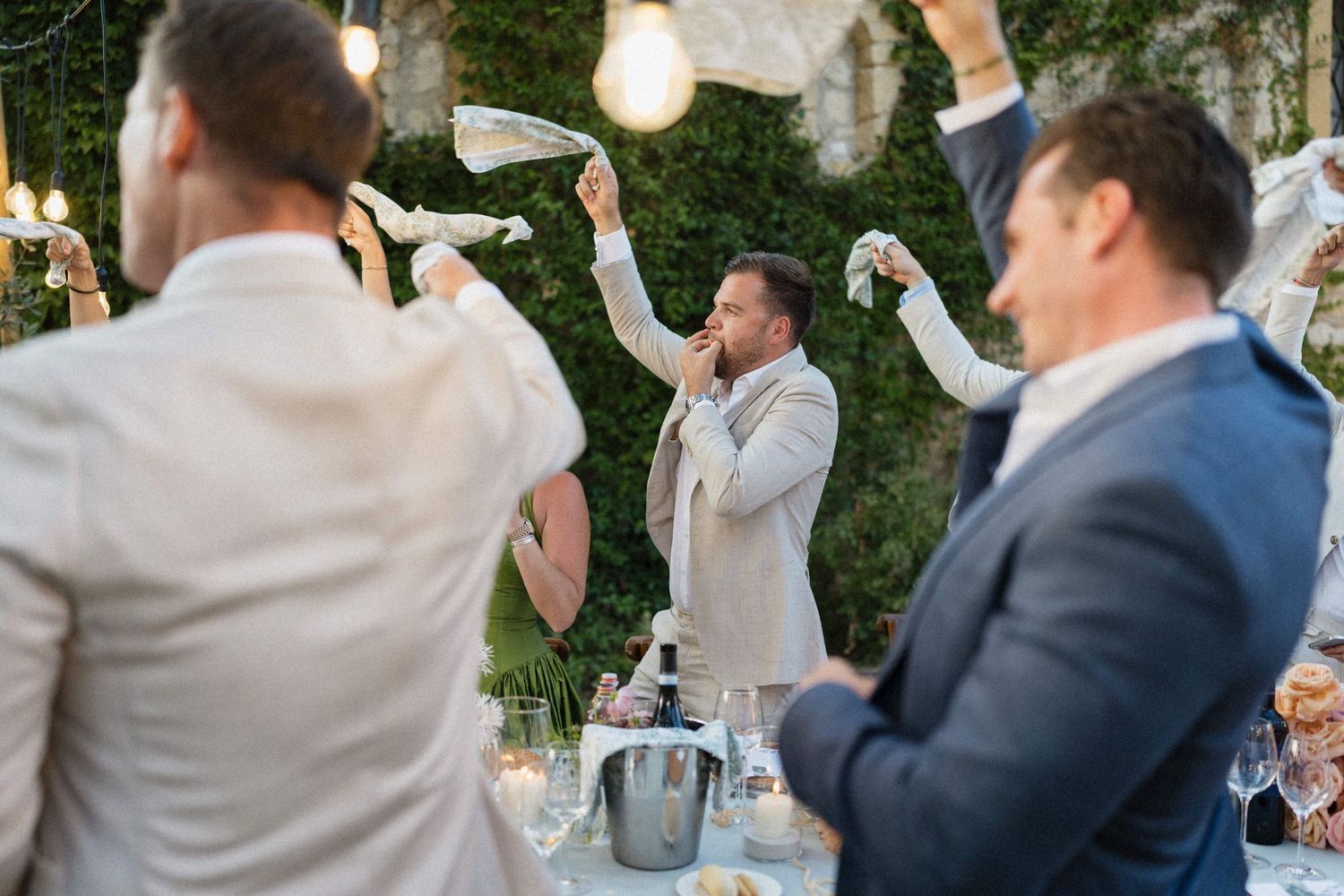 Men in suits waving white napkins at an outdoor wedding reception dinner party with champagne and string lights.
