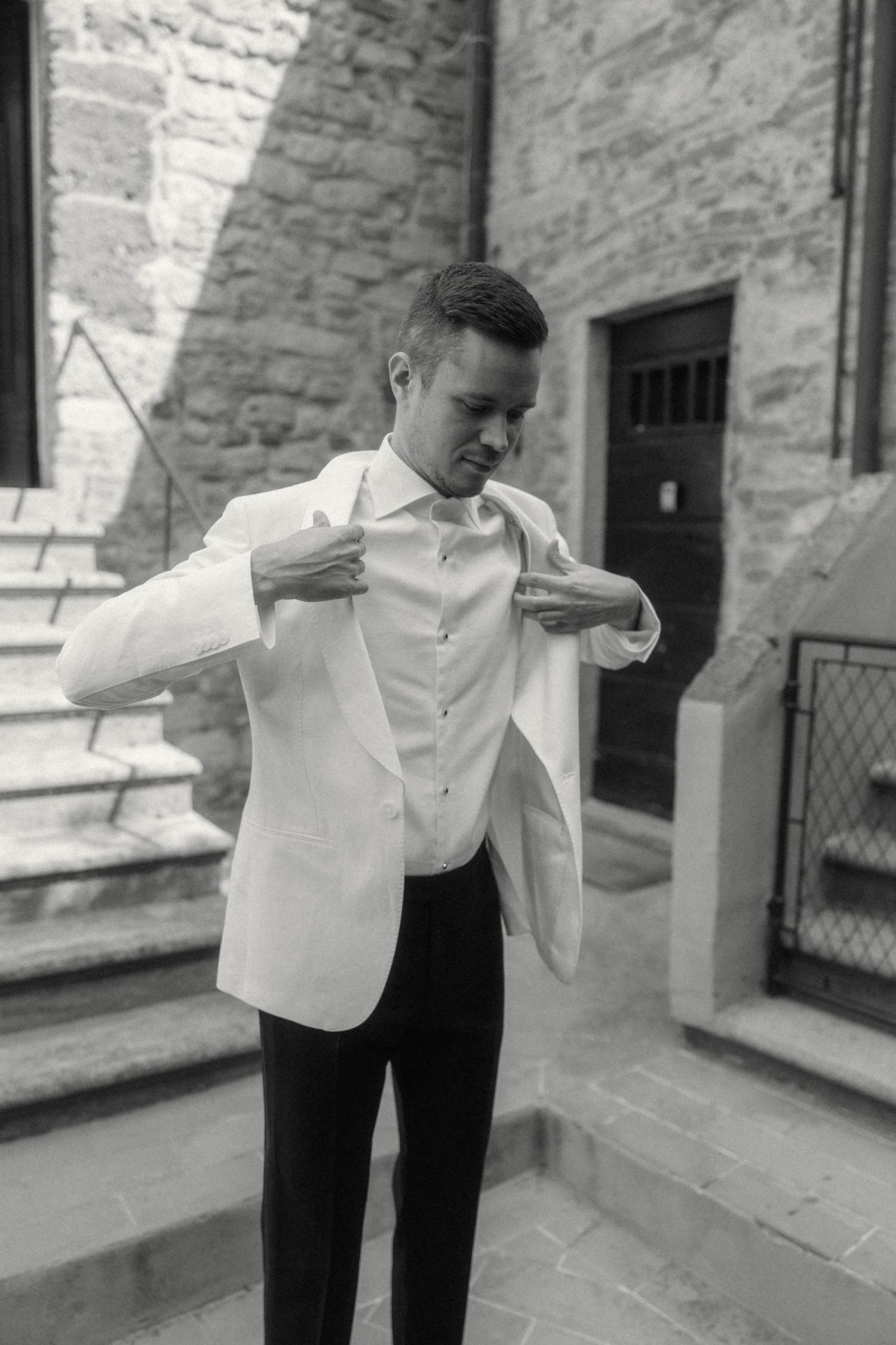 Young groom in white tuxedo jacket adjusting his lapels outdoors near stone building steps in black and white photo.