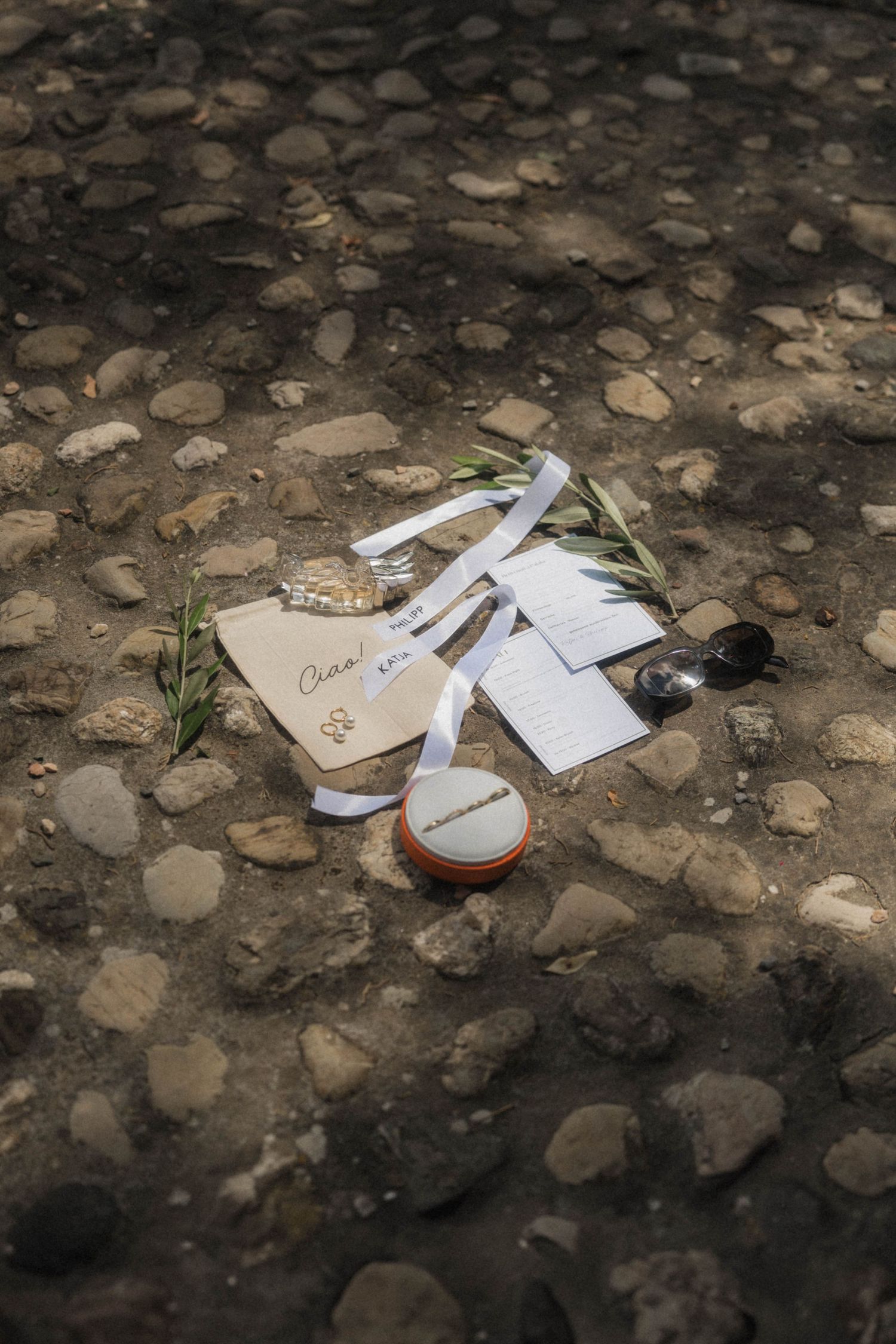 Wedding stationery, rings, and accessories arranged on rustic cobblestone ground at an outdoor ceremony.