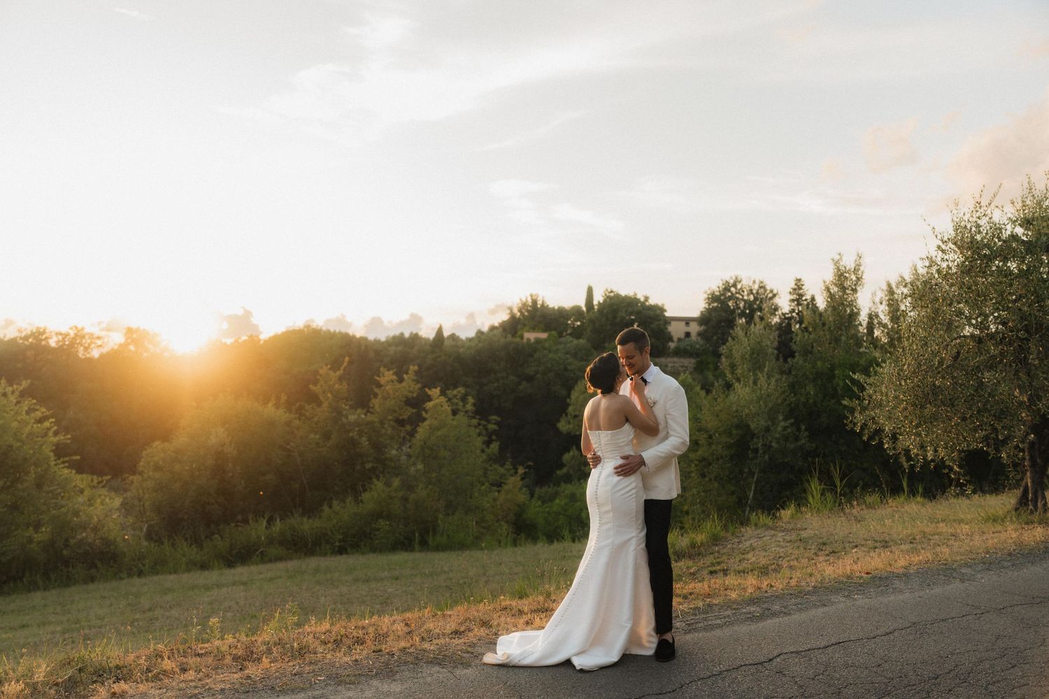 Bride and groom embracing on a road at golden sunset with lush green trees glowing in warm evening light.