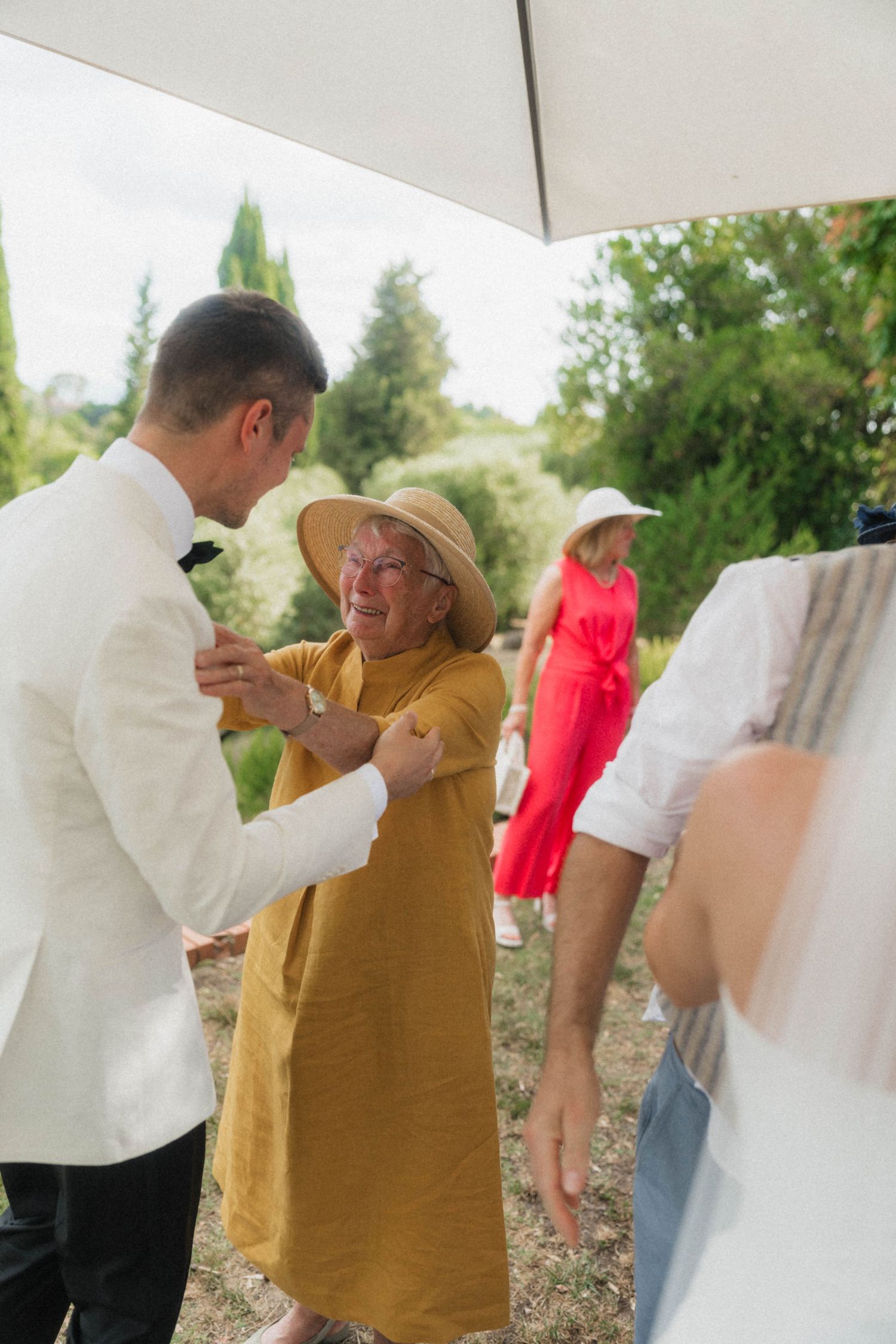 Groom in white jacket warmly greeting elderly female guest in mustard dress and hat at outdoor wedding.