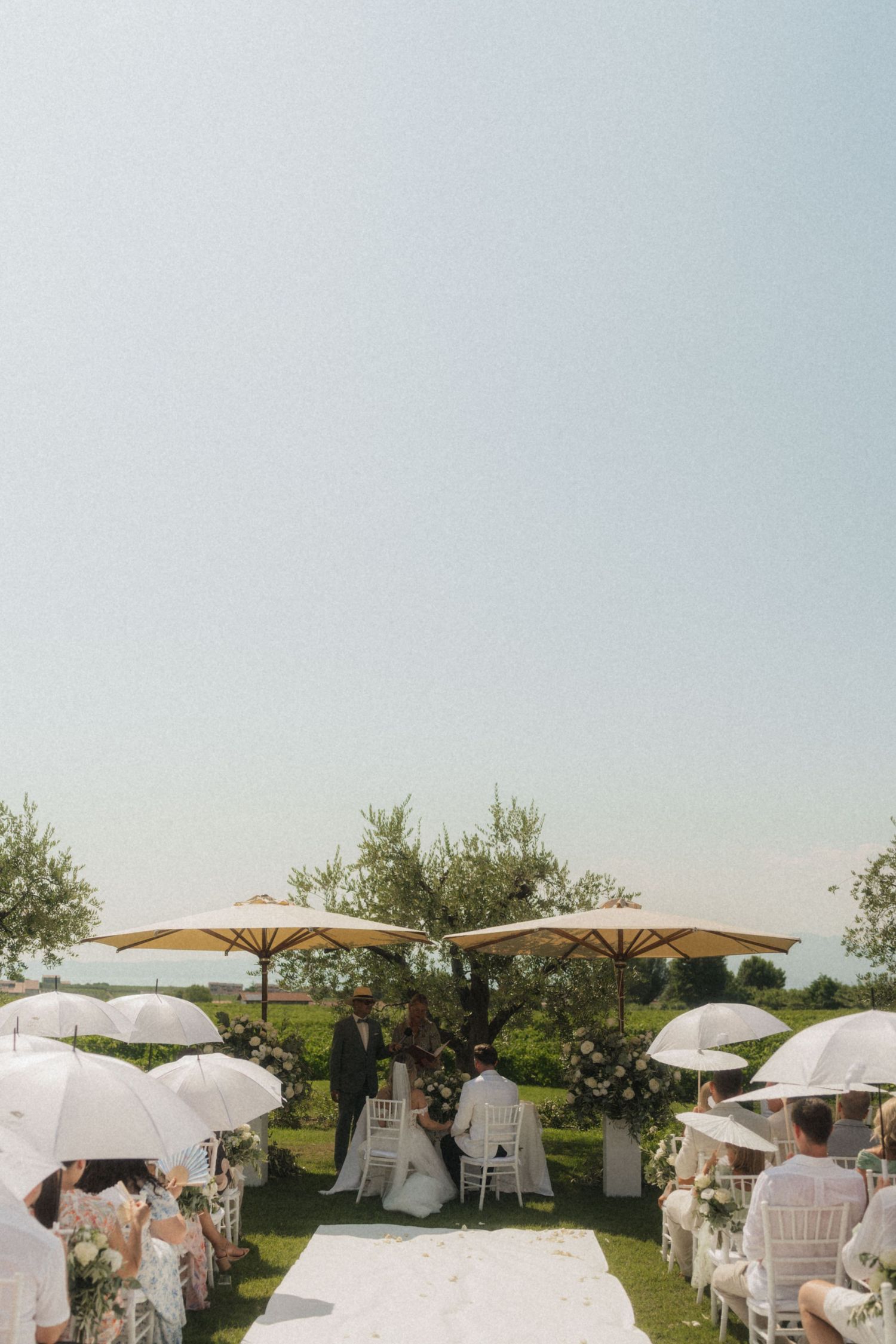 Elegant outdoor wedding ceremony with guests holding white parasols under large shade umbrellas.
