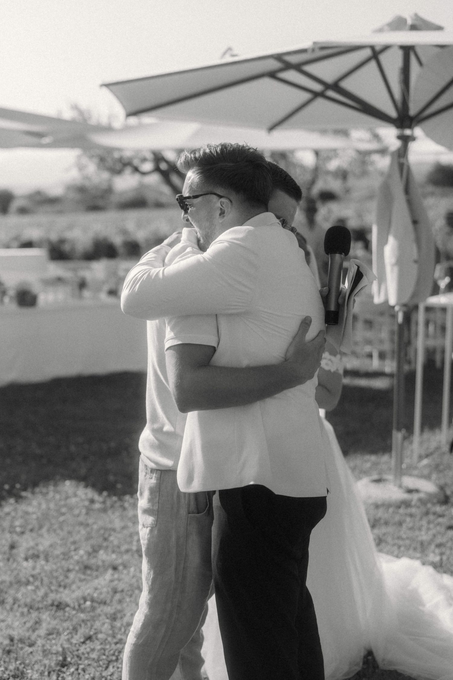 Black and white photo of groom sharing emotional hug with guest at outdoor wedding ceremony.