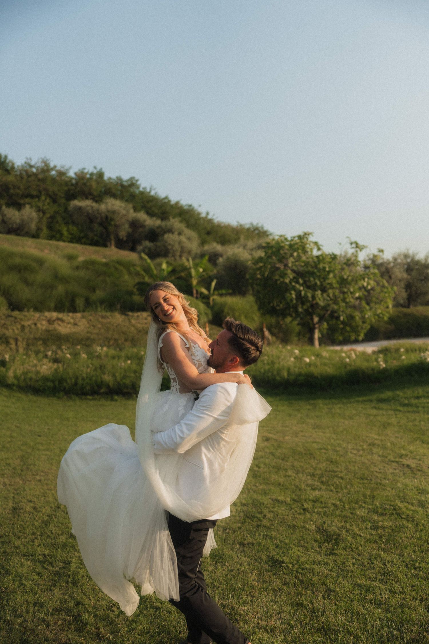 Groom lifting laughing bride in white gown during golden hour outdoor wedding portrait.