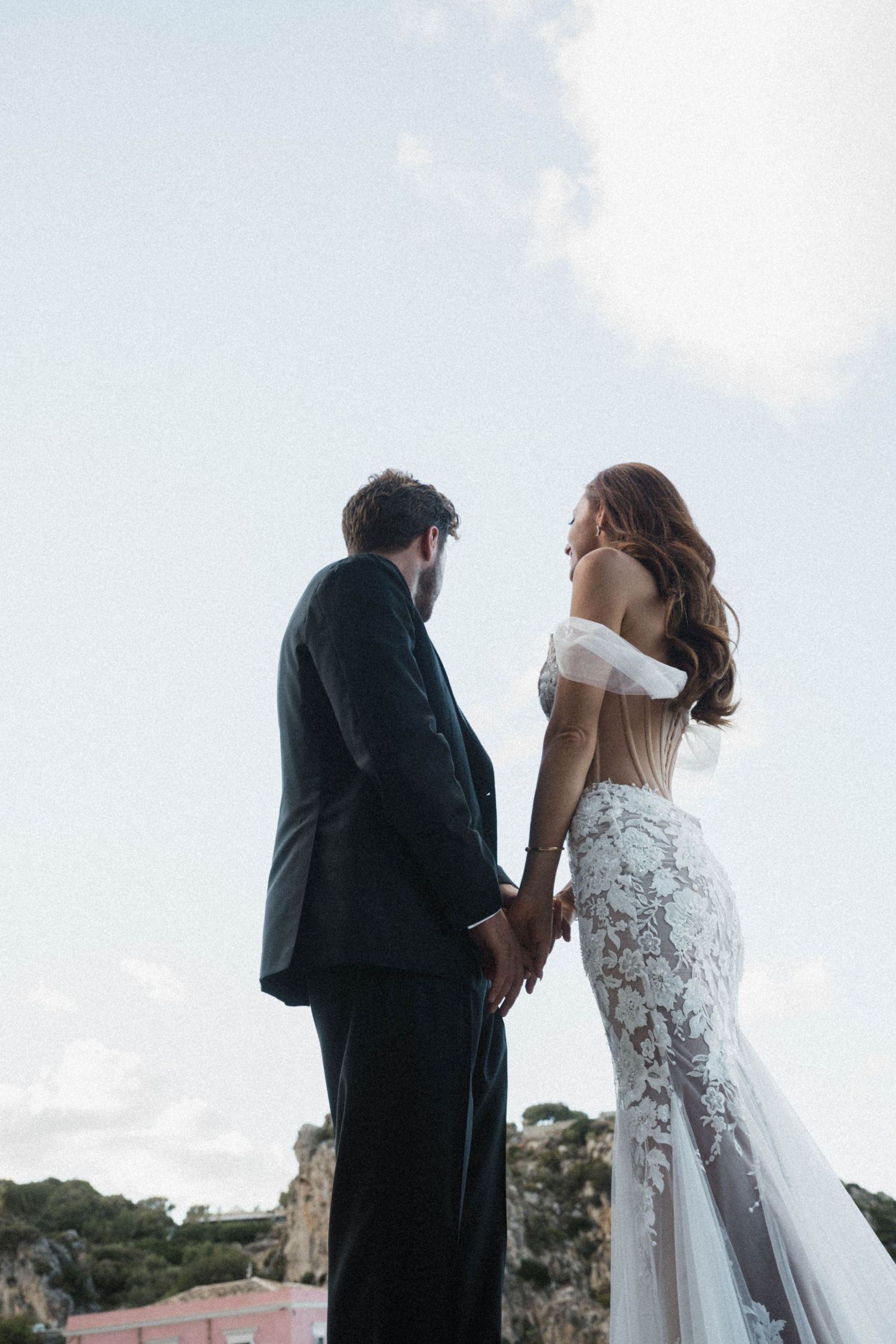 Bride and groom holding hands against bright sky, bride wearing elegant backless lace mermaid wedding dress.