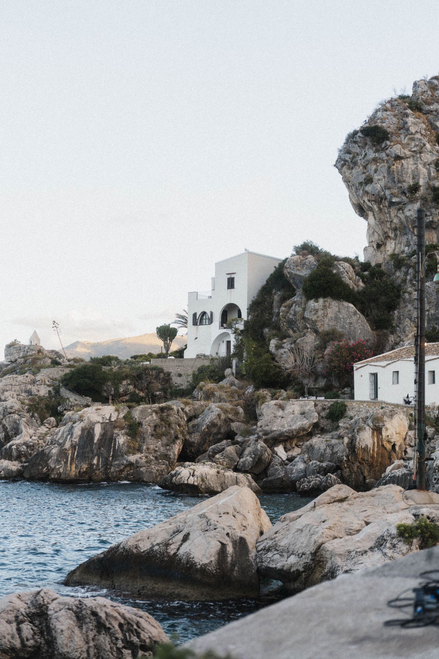 White Mediterranean villa perched on rocky coastal cliffs overlooking calm blue sea at golden hour.