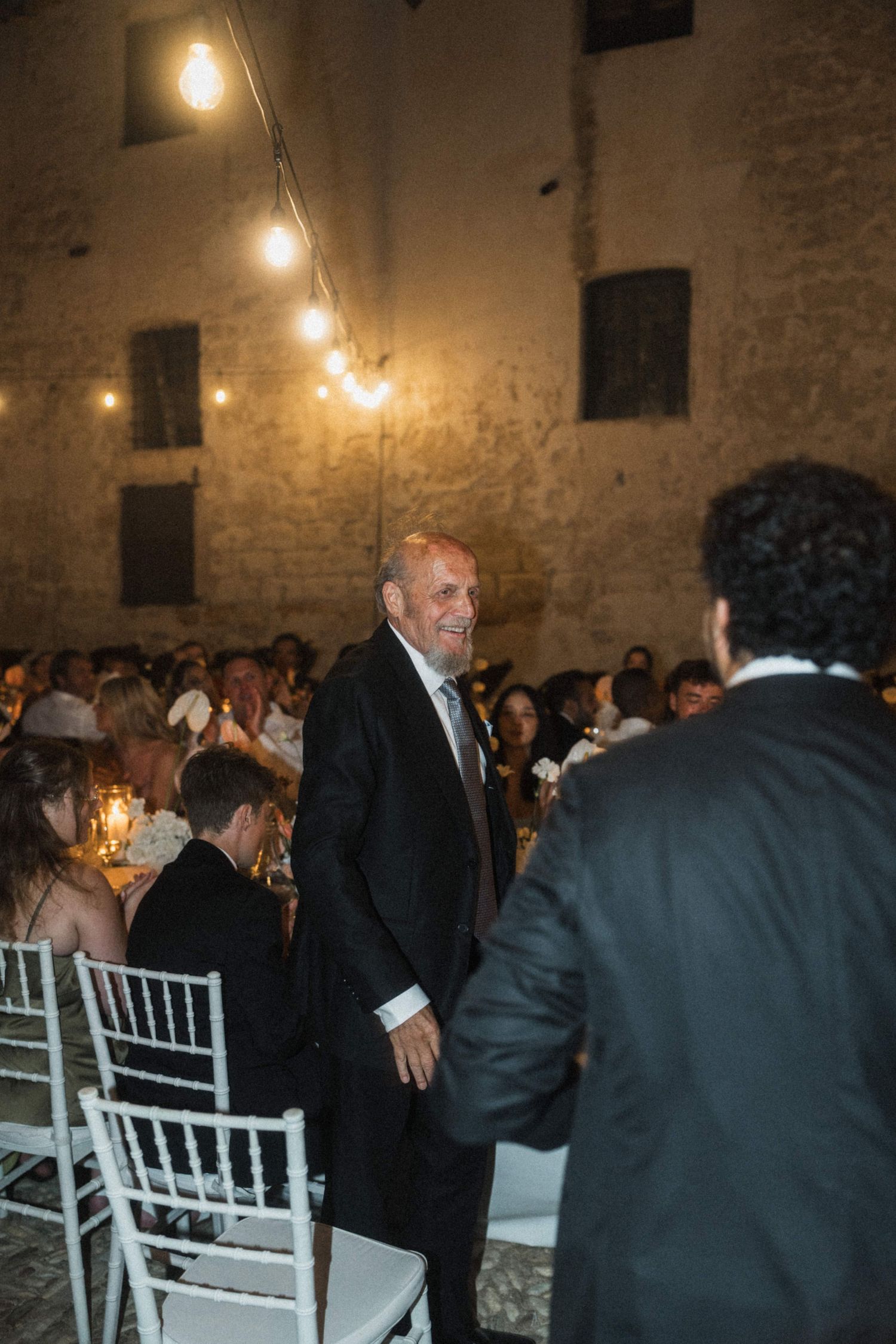 Elegantly dressed man in a dark suit smiling at guests during an indoor wedding reception with string lights.