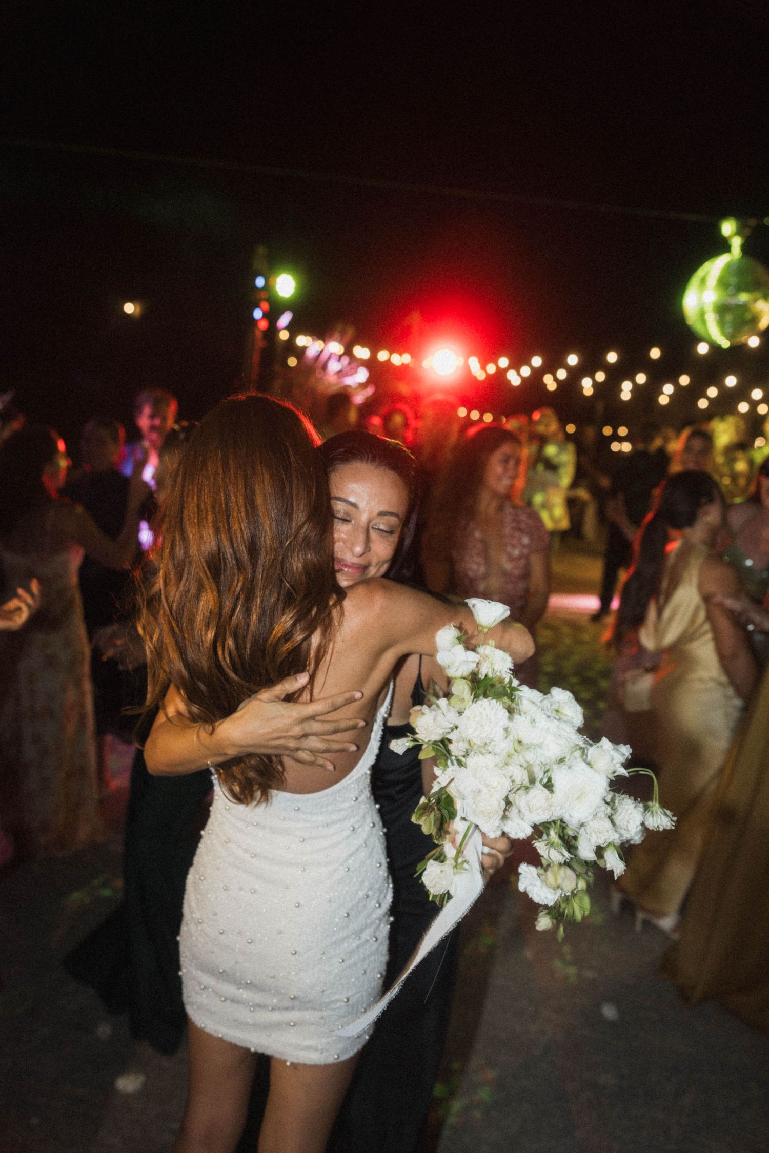 Bride in white dress hugging guest while holding white rose bouquet at outdoor wedding reception.
