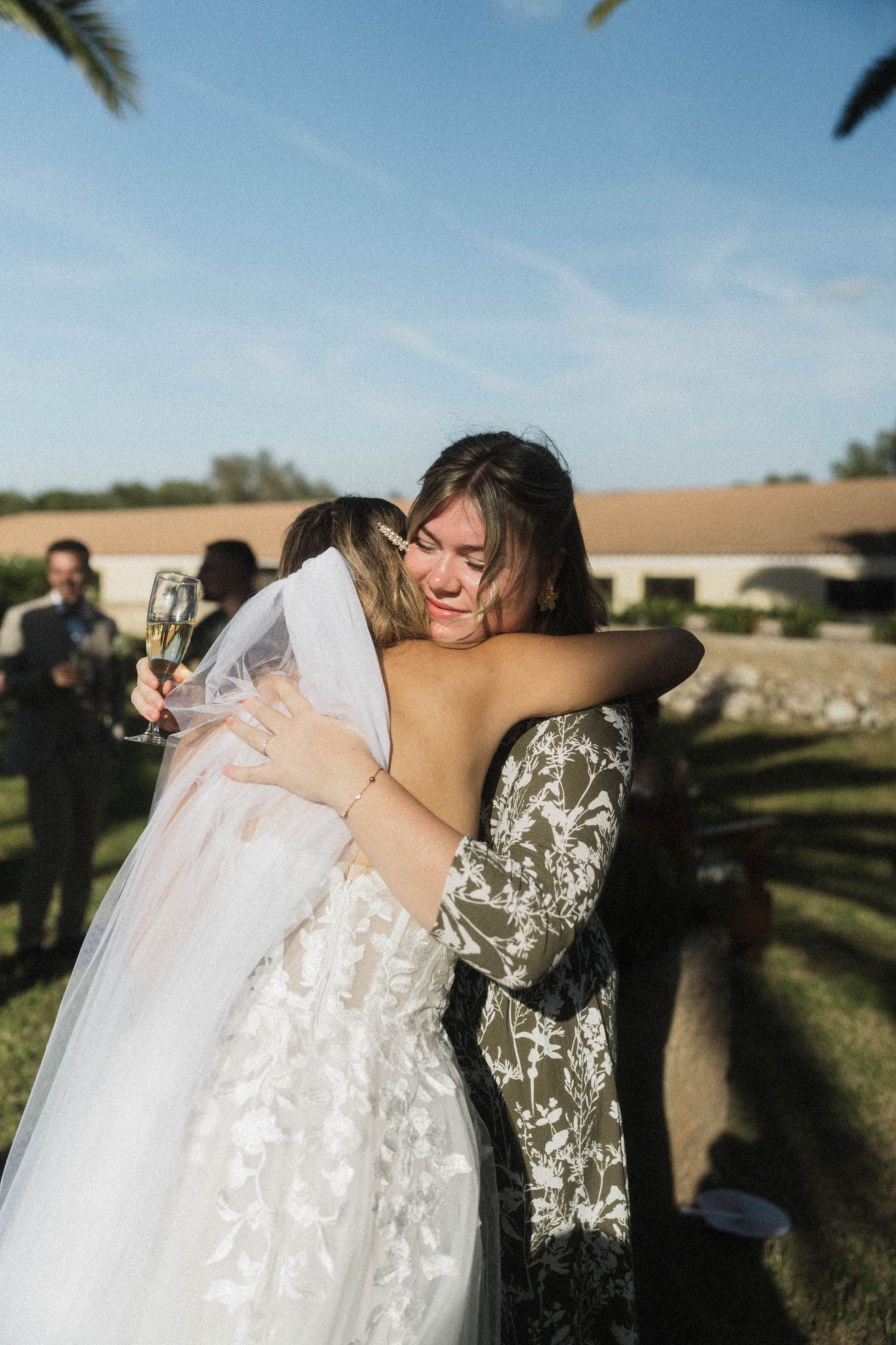 Emotional wedding guest hugs bride in lace gown while holding champagne at outdoor celebration.