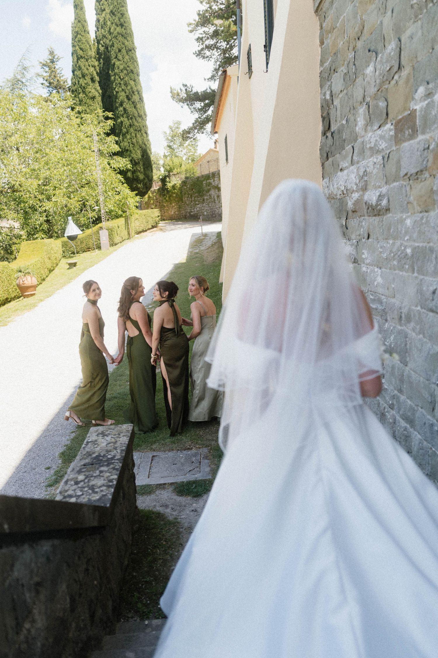 Bride in white veil and gown approaching four bridesmaids in olive green dresses outdoors.