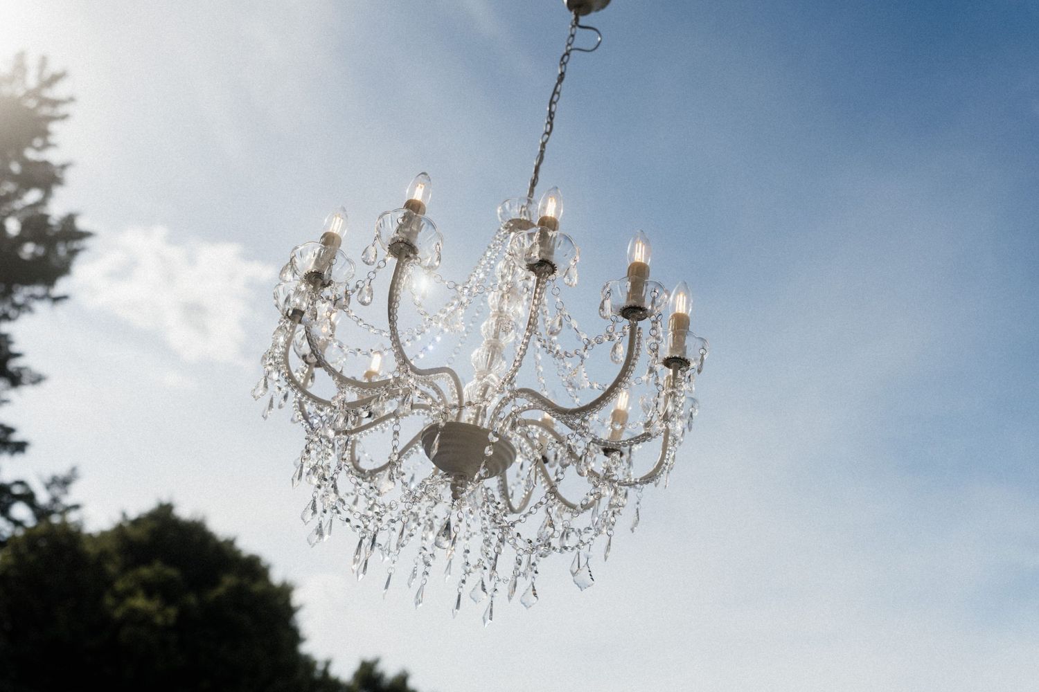 Ornate crystal chandelier with candle lights hanging outdoors against a blue sky backdrop.