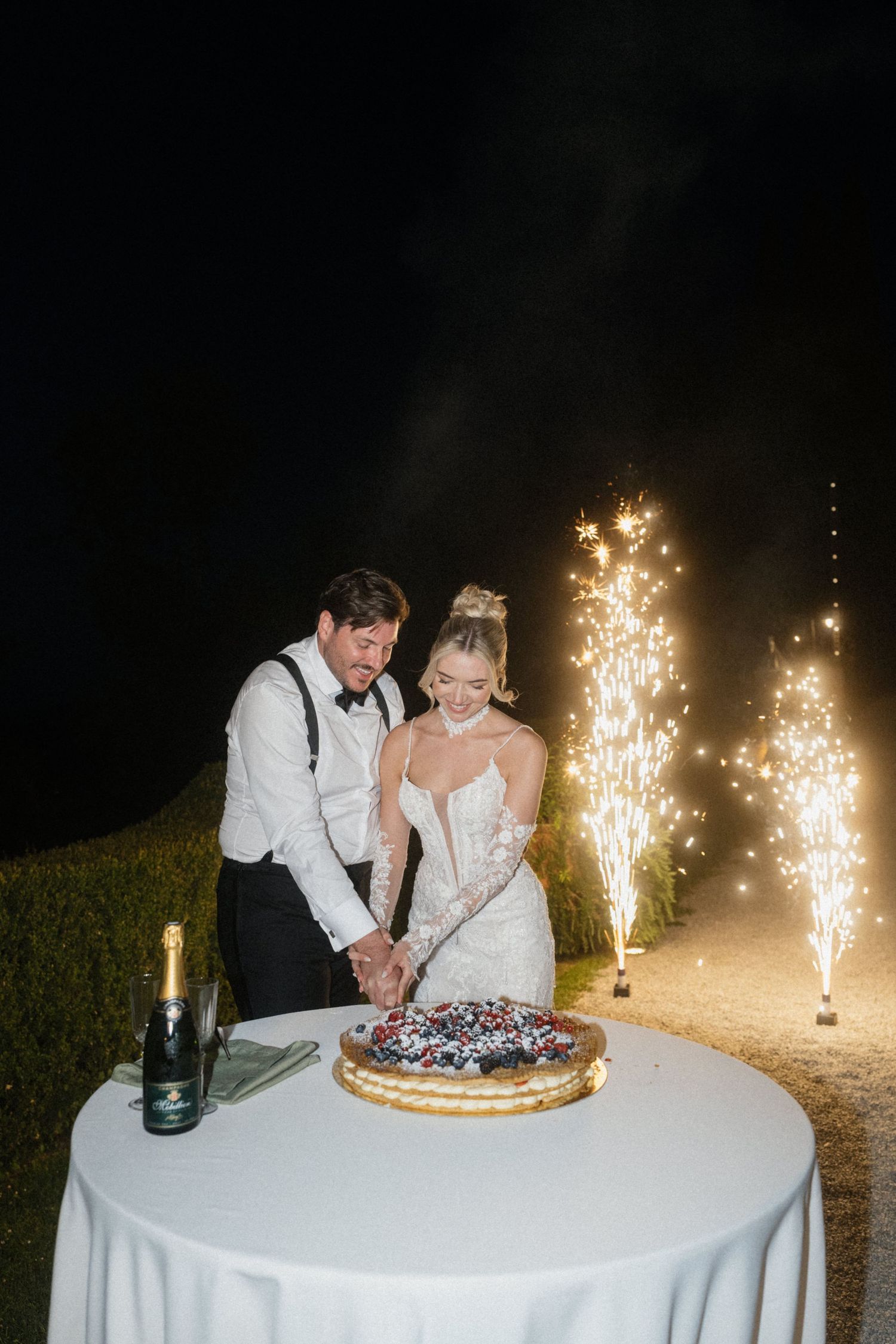 Bride and groom cutting wedding cake outdoors at night with sparkler fireworks display behind them.