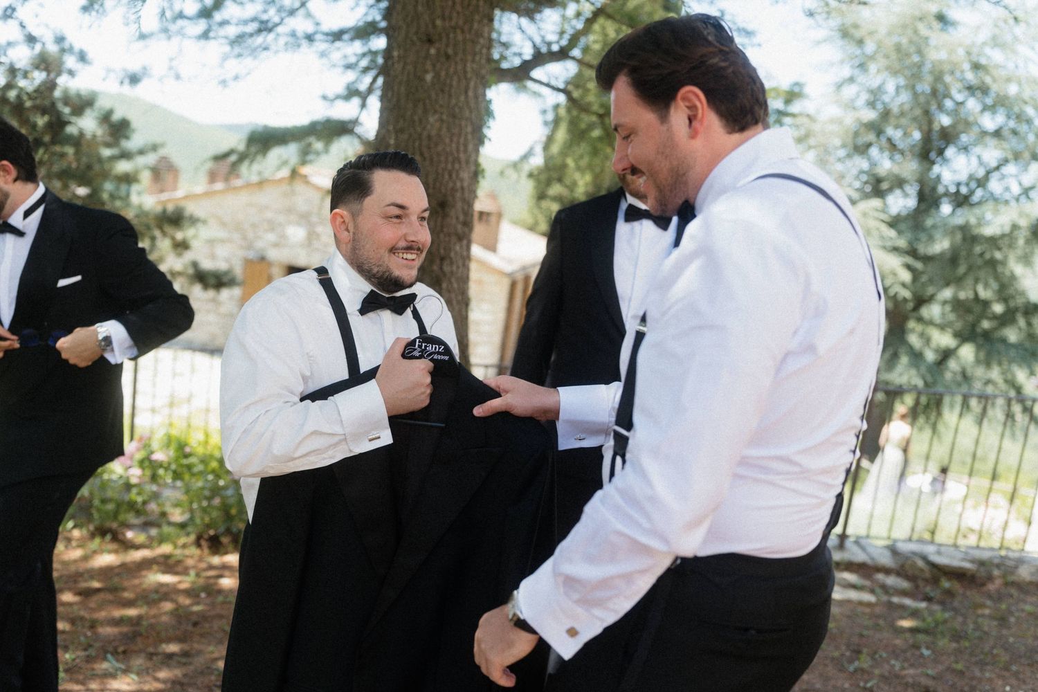 Two groomsmen in black tuxedos and bow ties laughing while helping each other dress outdoors.
