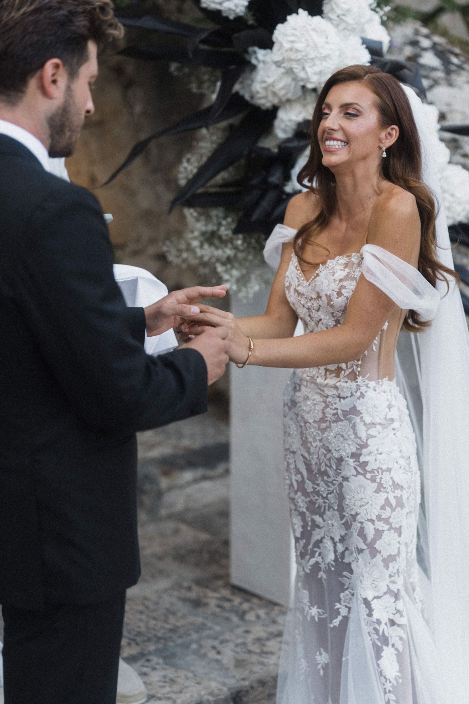 Smiling bride in lace gown exchanges rings with groom at outdoor wedding altar.