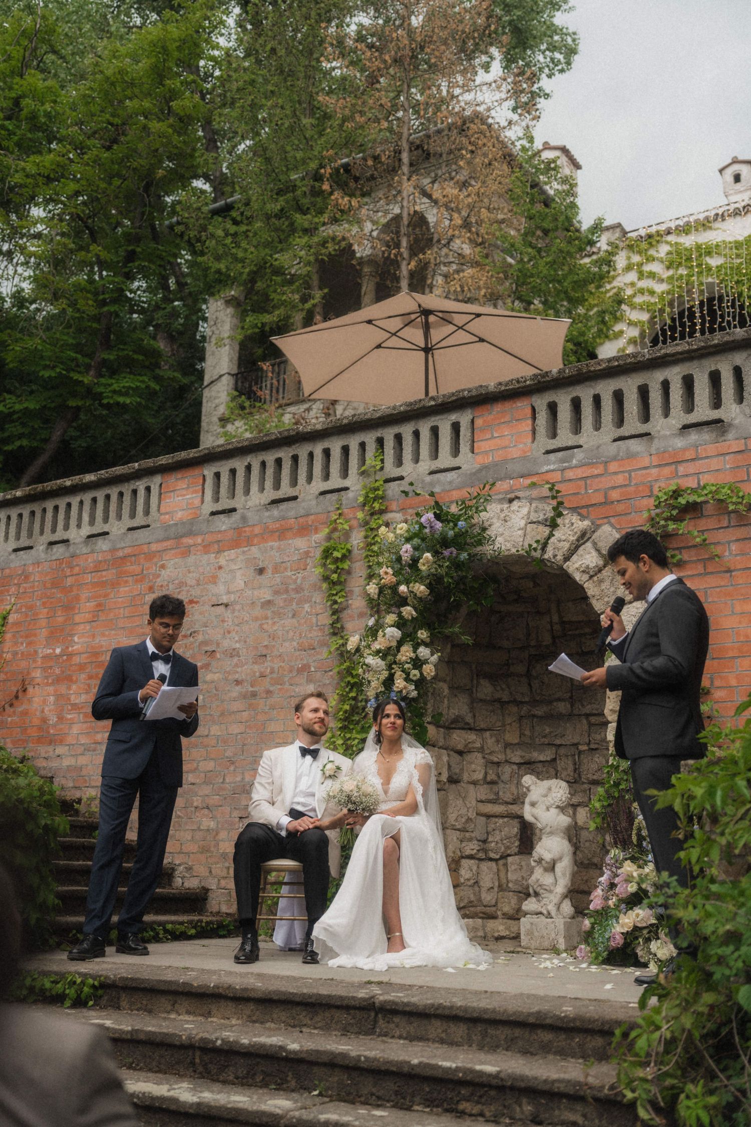 Bride and groom seated at floral arch altar during outdoor wedding ceremony at rustic villa.