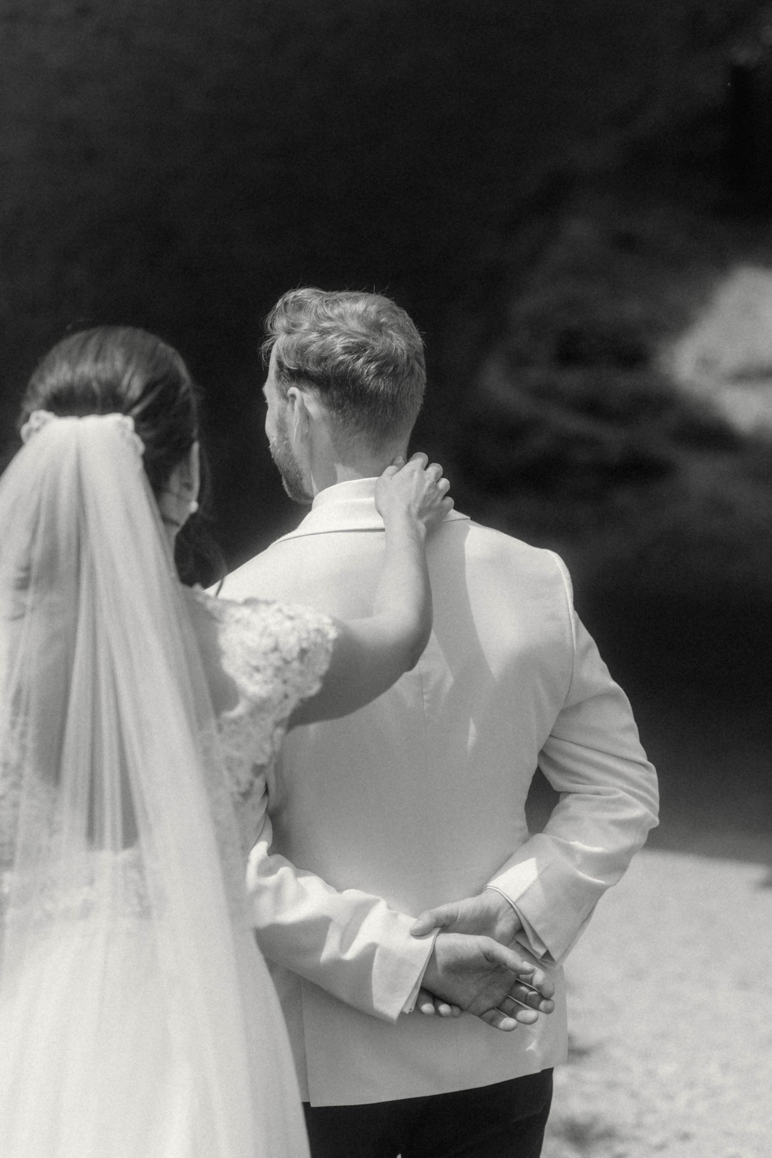 Bride and groom standing together from behind, bride in lace gown with veil, groom in white jacket.