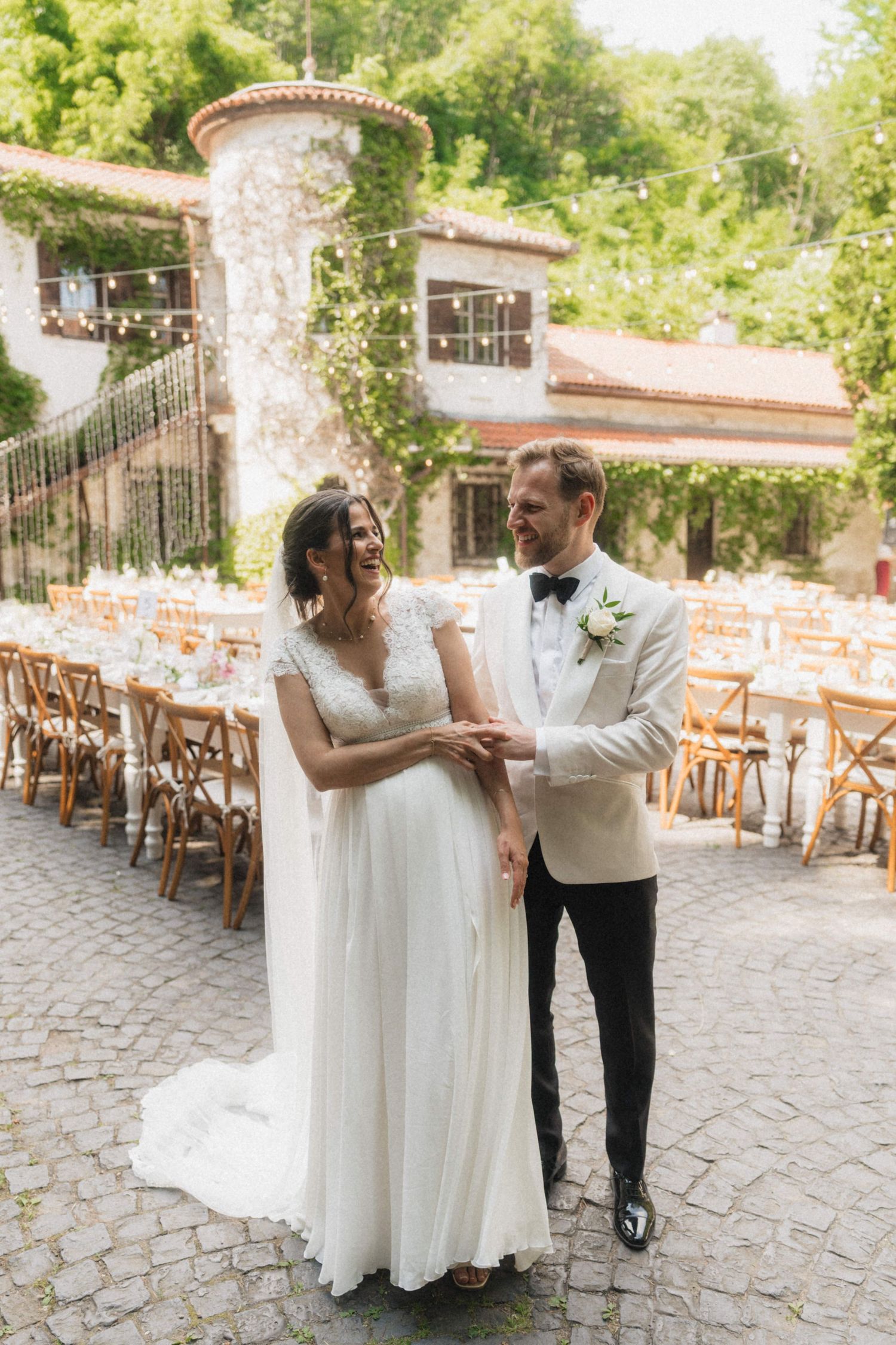 Smiling bride and groom in white lace dress and ivory tuxedo posing at an outdoor stone wedding venue.