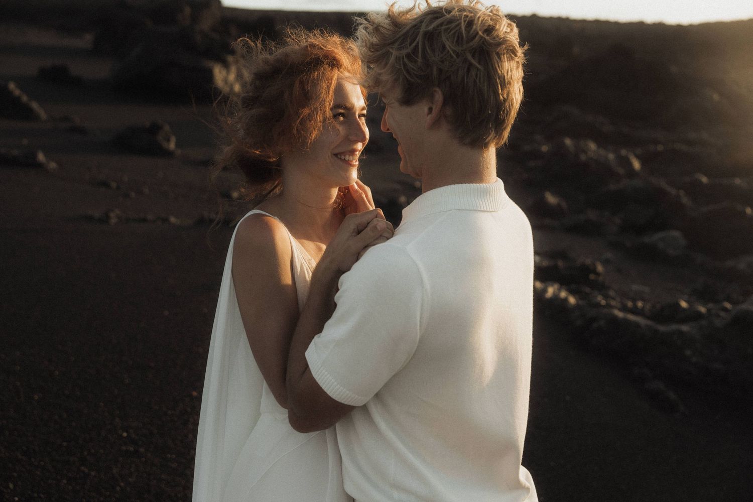 Bride and groom sharing romantic moment on black sand beach at golden sunset.