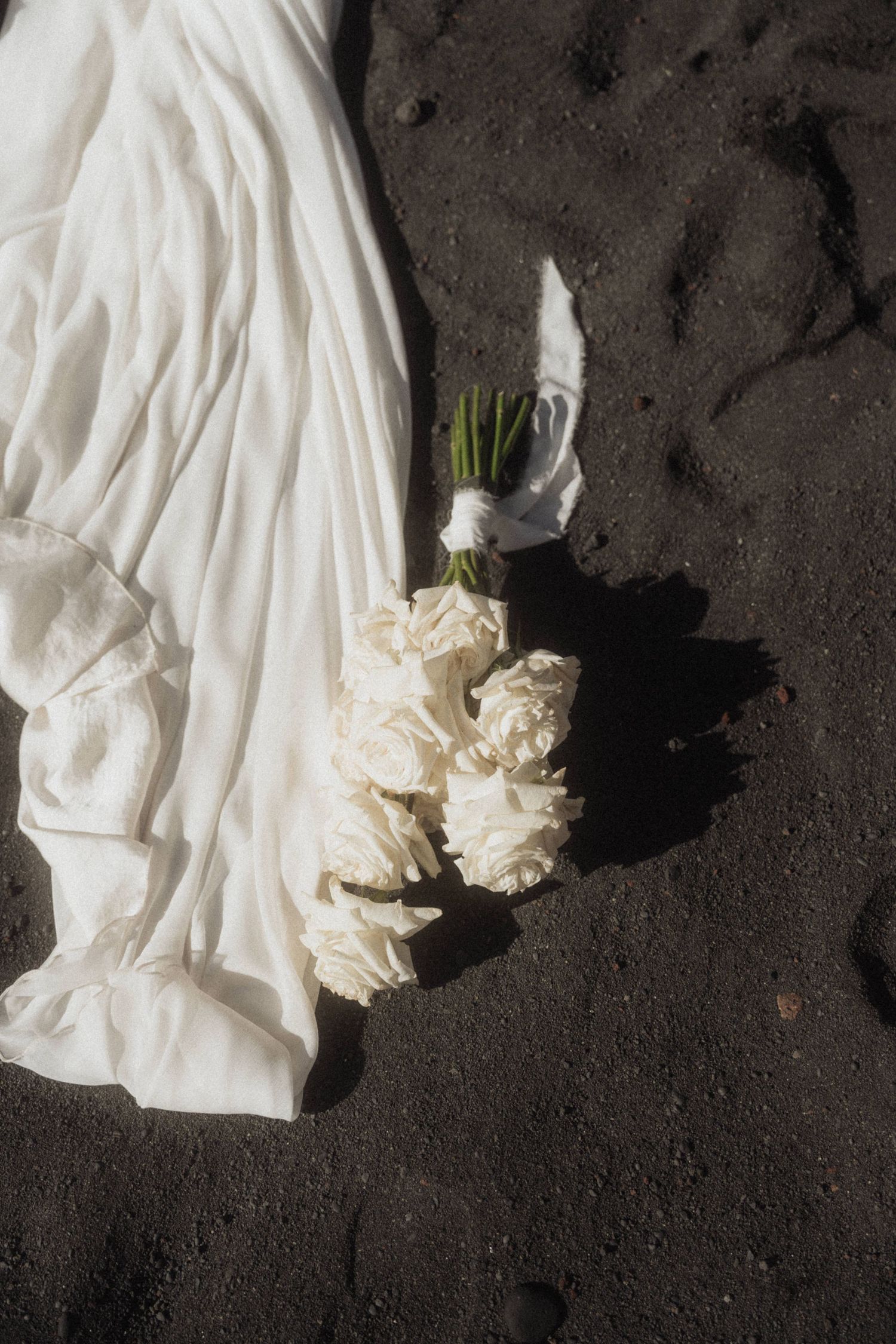 White bridal bouquet of roses resting on dark volcanic sand beside wedding dress train.