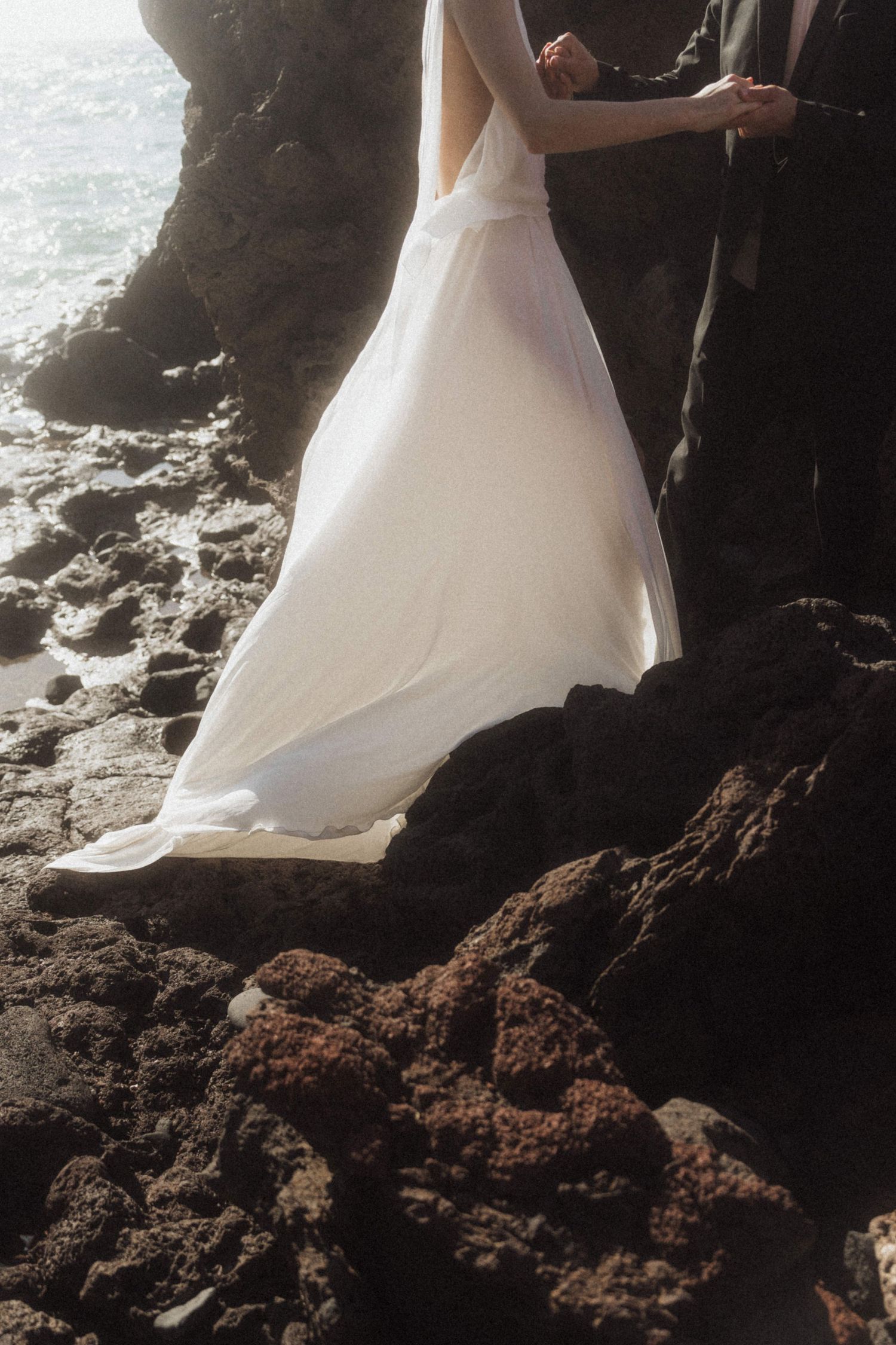 Bride in flowing white gown standing on volcanic rocks by ocean with groom.