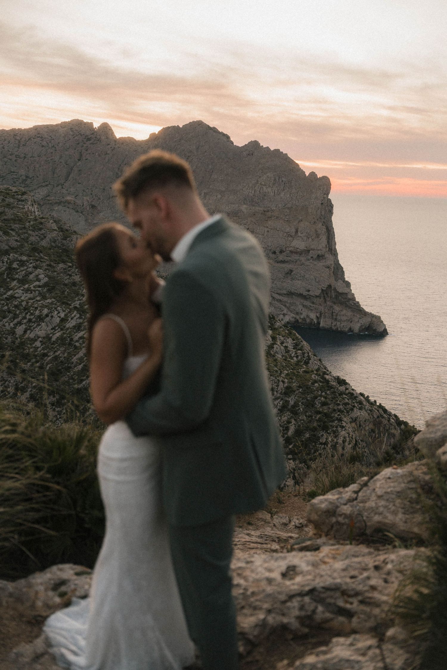 Couple shares a romantic kiss at sunset on a cliff overlooking dramatic rocky coastline and glowing orange sky.