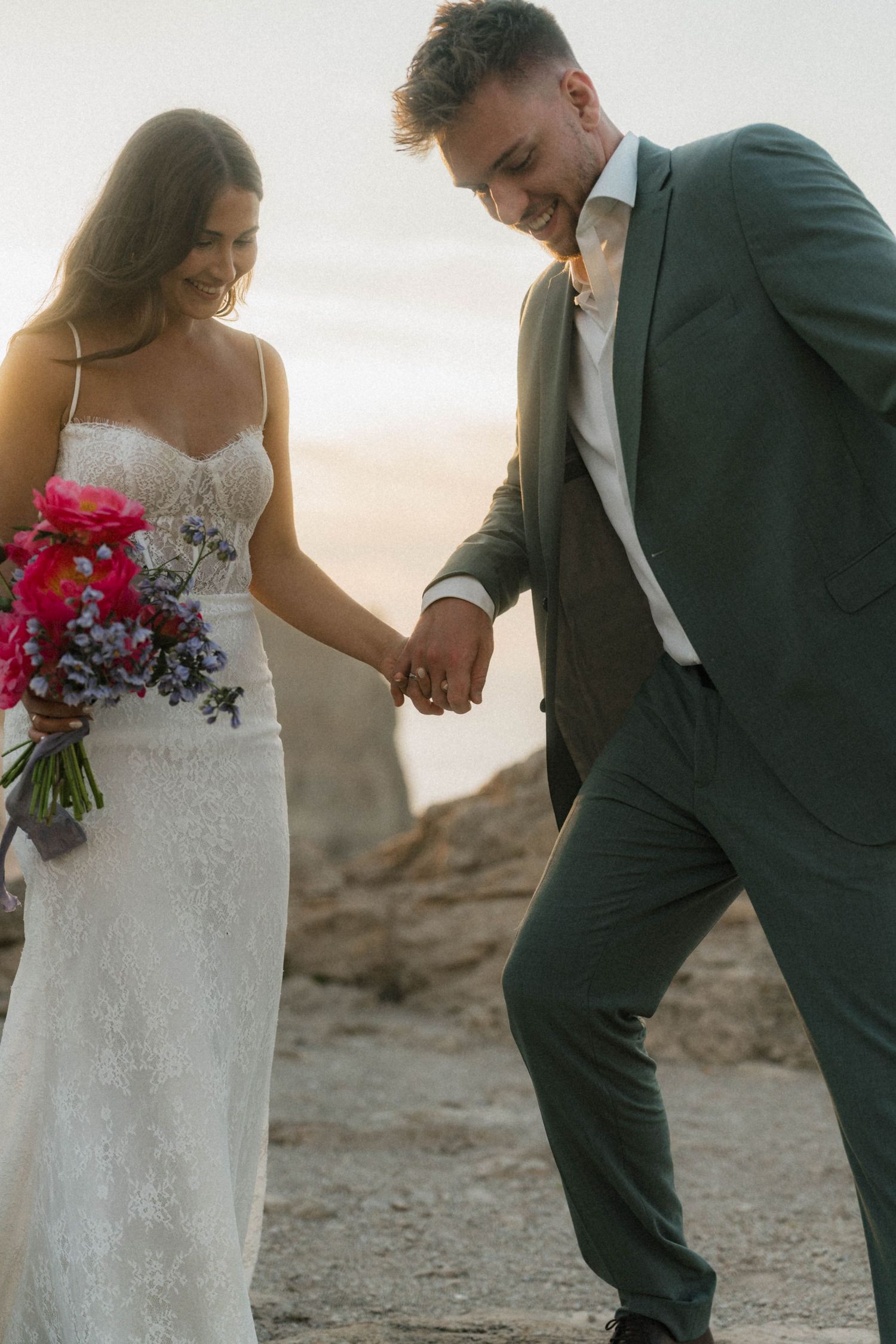 Smiling groom leads bride by hand across rocky terrain at sunset, bride in lace gown carrying colorful floral bouquet.