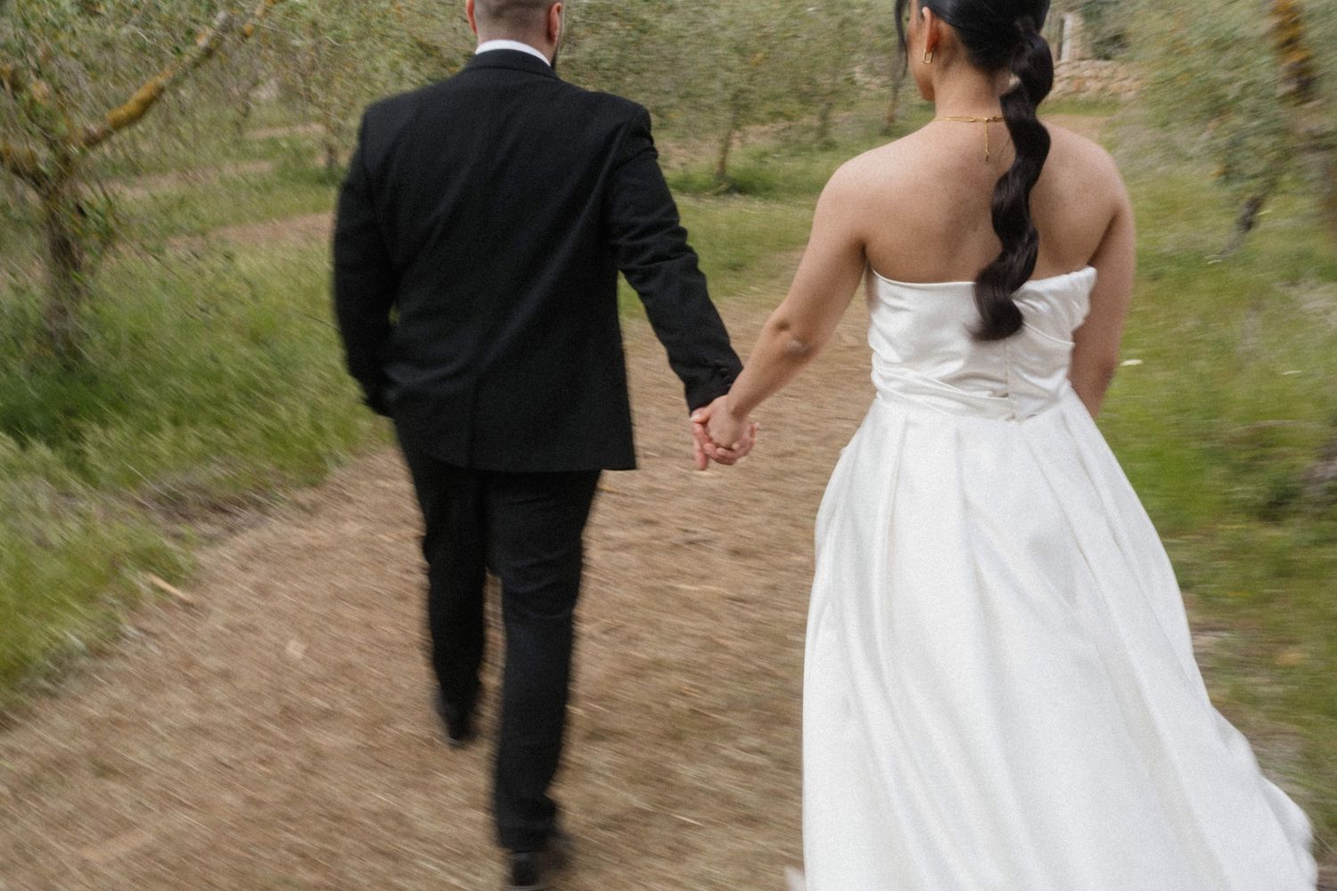 Bride and groom walking hand in hand down a rustic dirt path surrounded by olive trees.