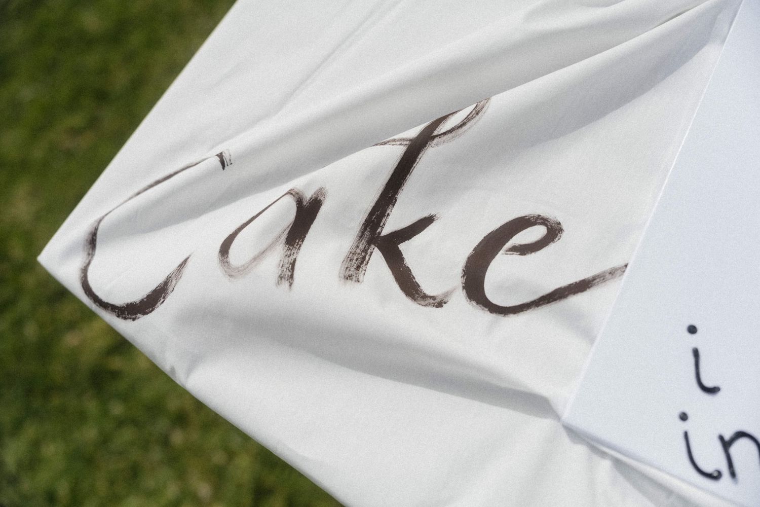 White wedding sign with cursive text reading Cake written in brown ink on grass background.