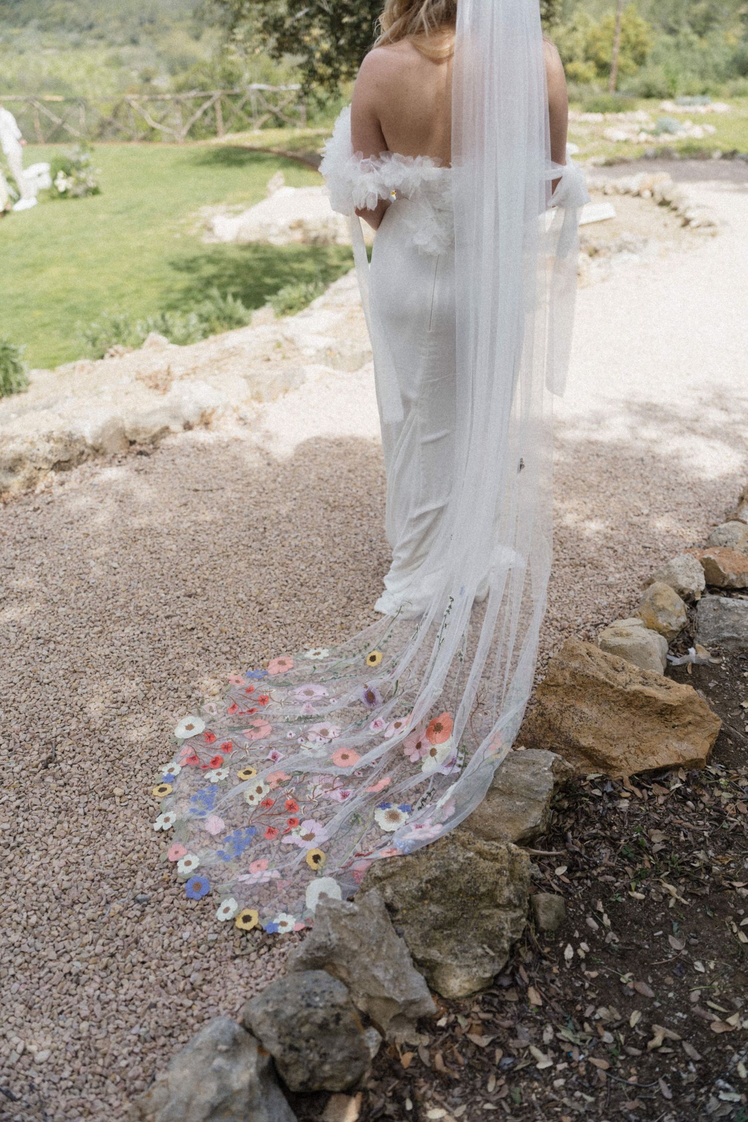 Bride in white backless gown with floral embroidered cathedral veil trailing on stone path outdoors.
