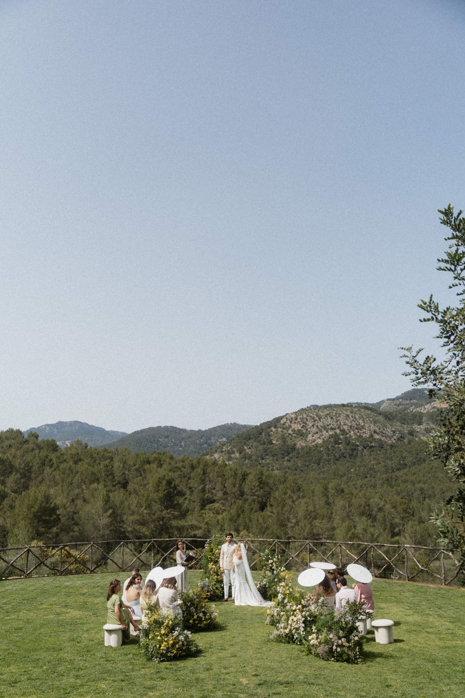 Outdoor wedding ceremony on a green lawn with mountain views, floral arrangements, and guests under parasols.