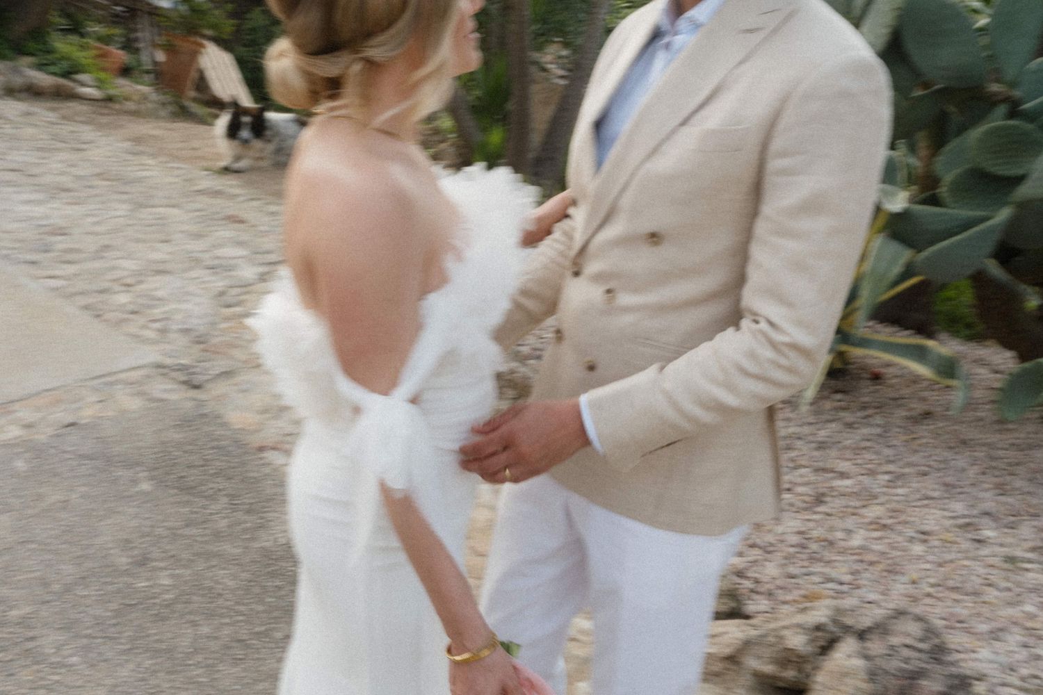 Bride in a backless white gown with feather wrap standing with groom in beige suit at an outdoor wedding venue.
