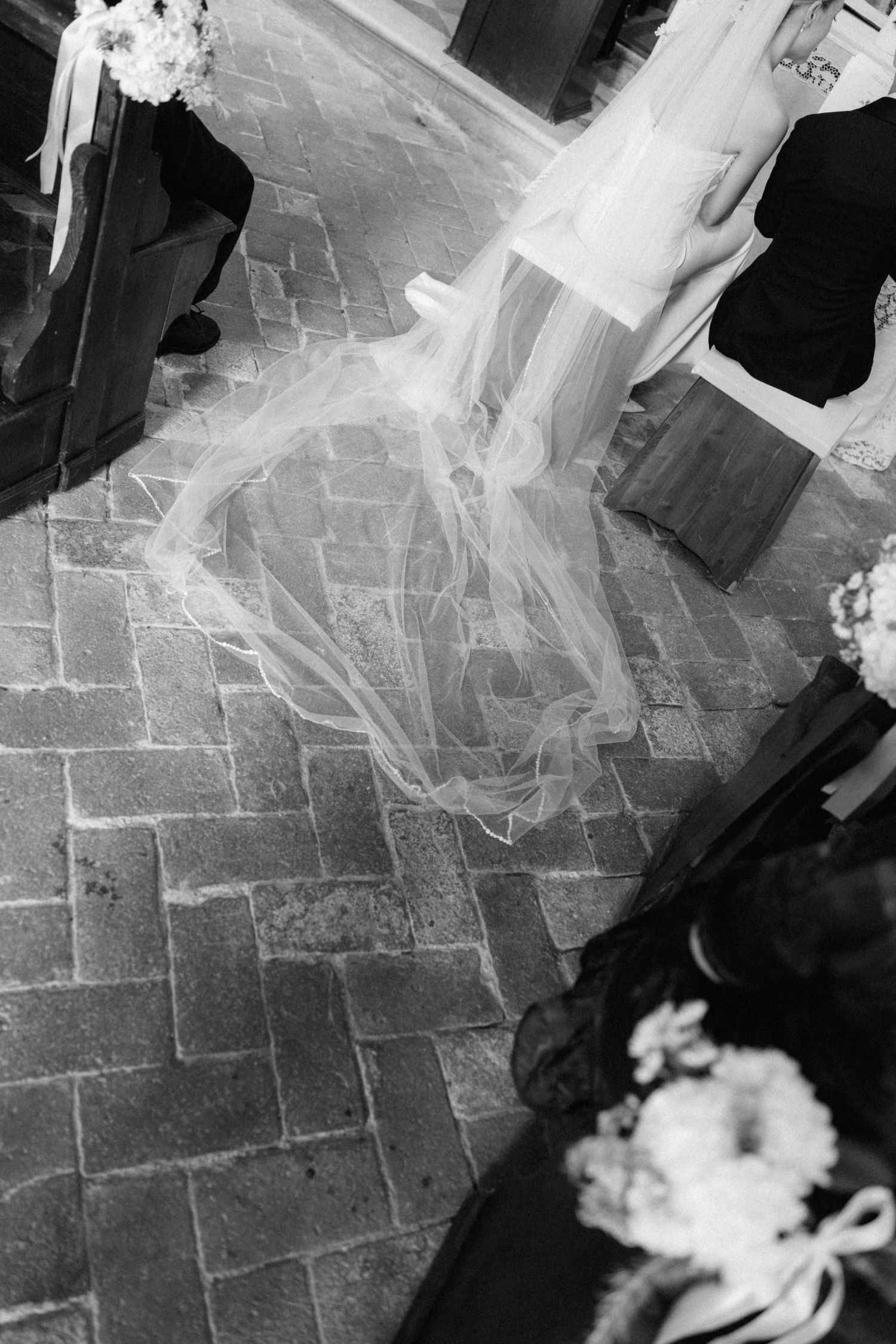 Black and white photo of a wedding veil trailing on herringbone brick floor during ceremony.